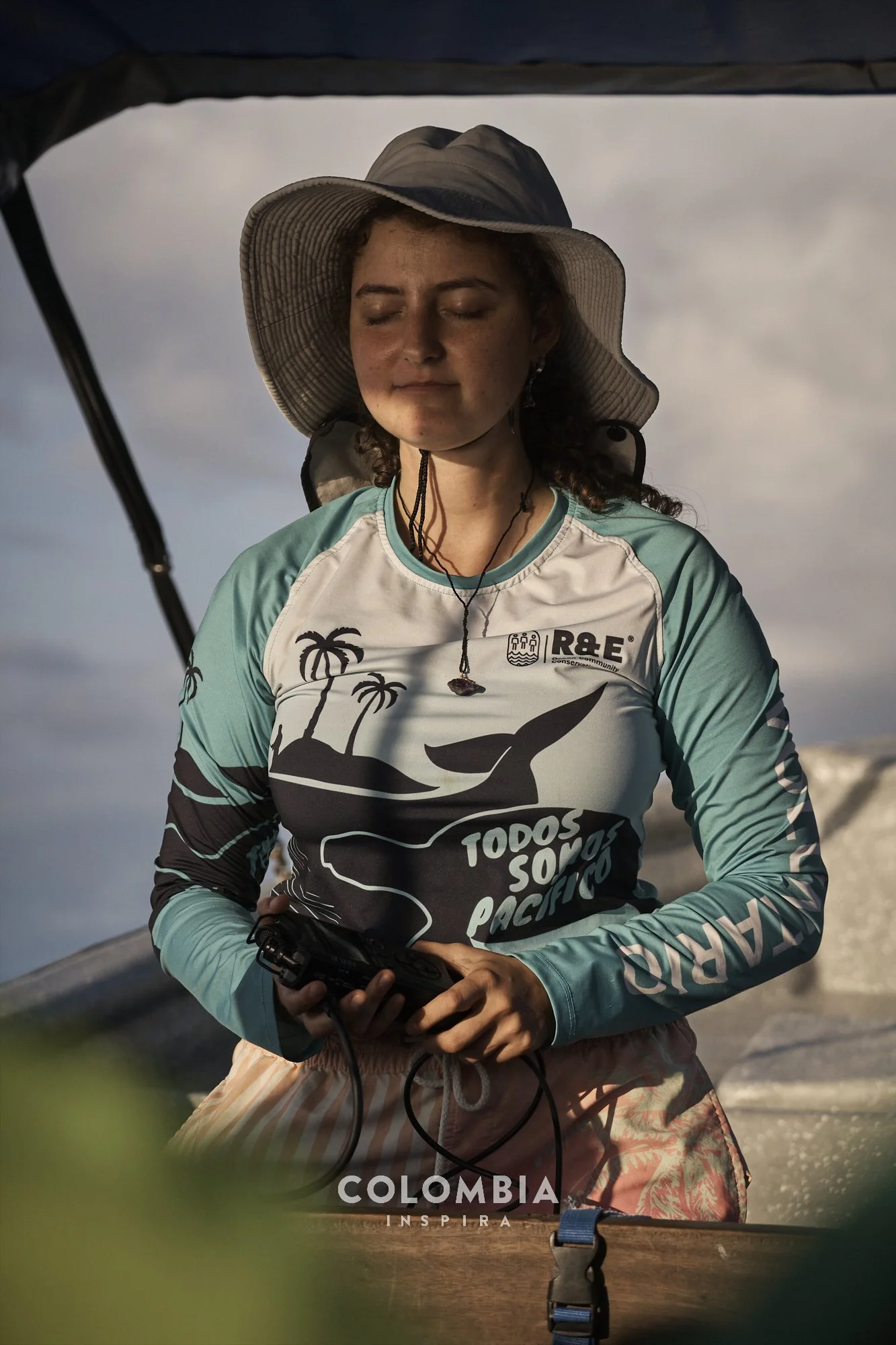 Young woman with curly hair wearing a wide-brimmed hat, teal and white long-sleeve shirt with palm tree and whale graphics, holding a device, outdoors under cloudy sky in Colombia.