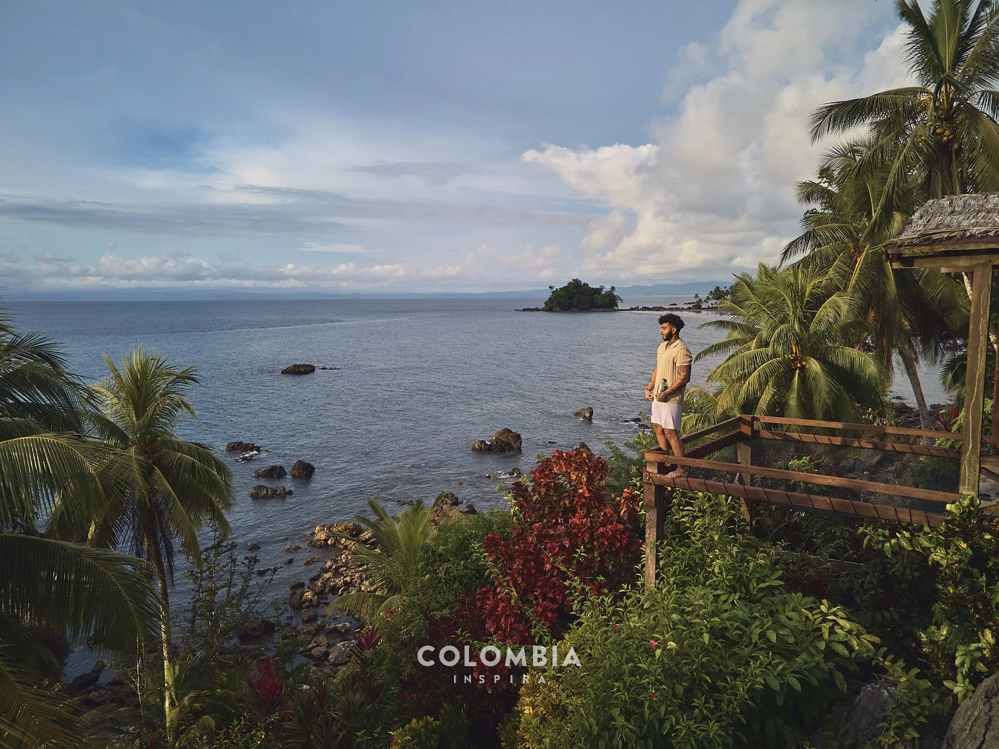 A man standing on a wooden balcony overlooking the ocean with lush green palm trees and colorful plants nearby, under a partly cloudy sky, in Colombia.