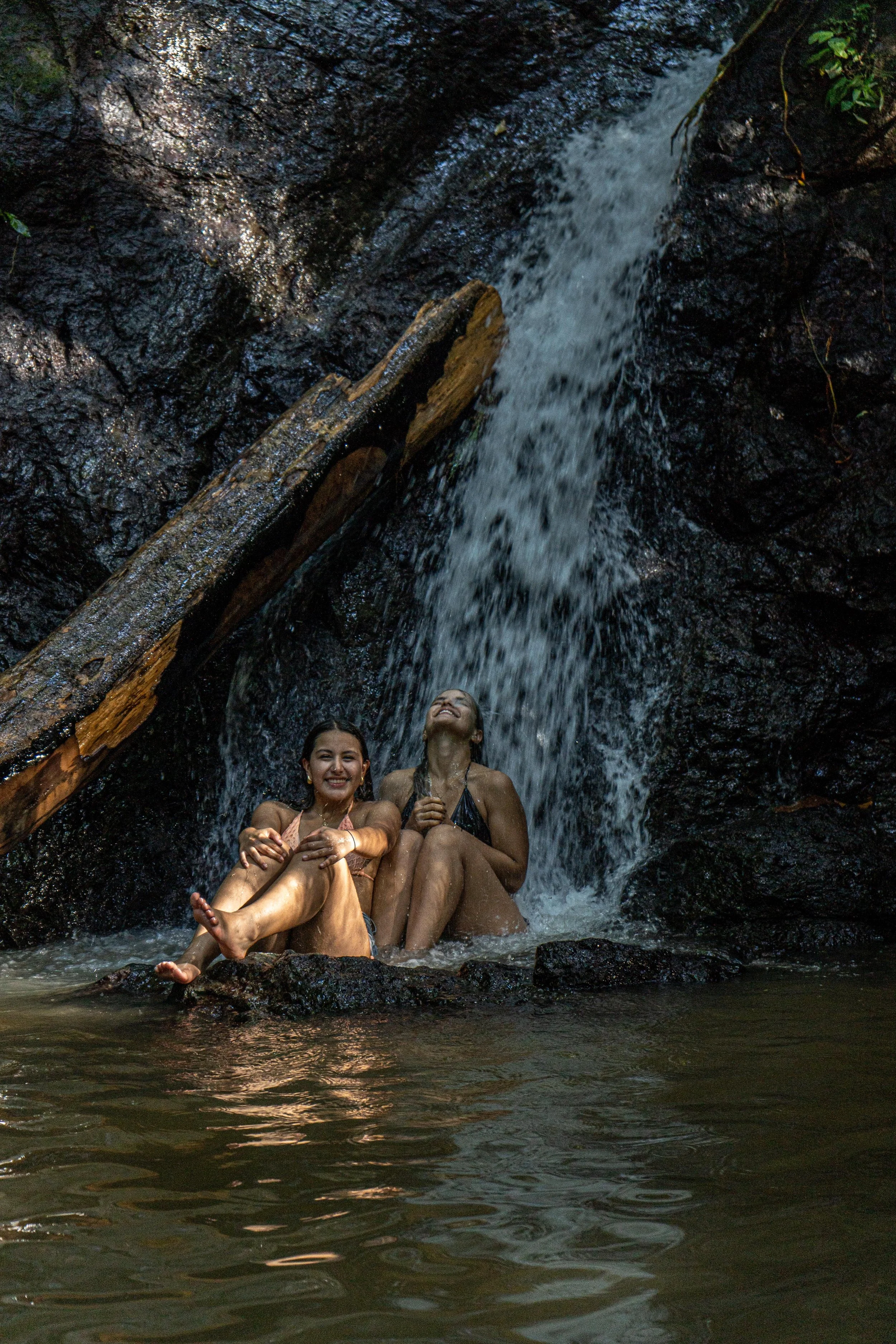 Two women sitting in a small pool under a waterfall in a lush, tropical environment.