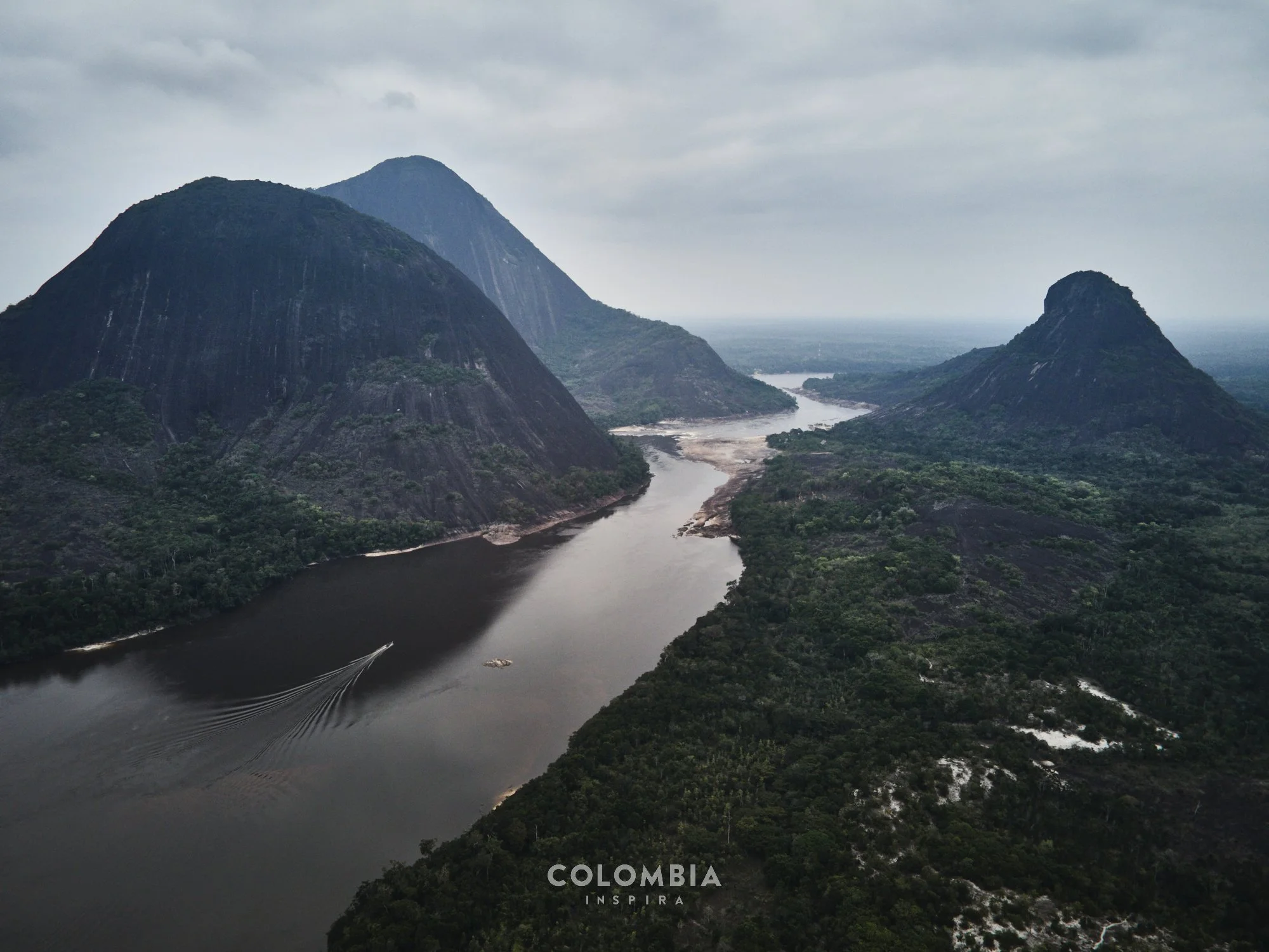 Aerial view of a river flowing through lush green mountains in Colombia, with steep slopes and cloudy sky.