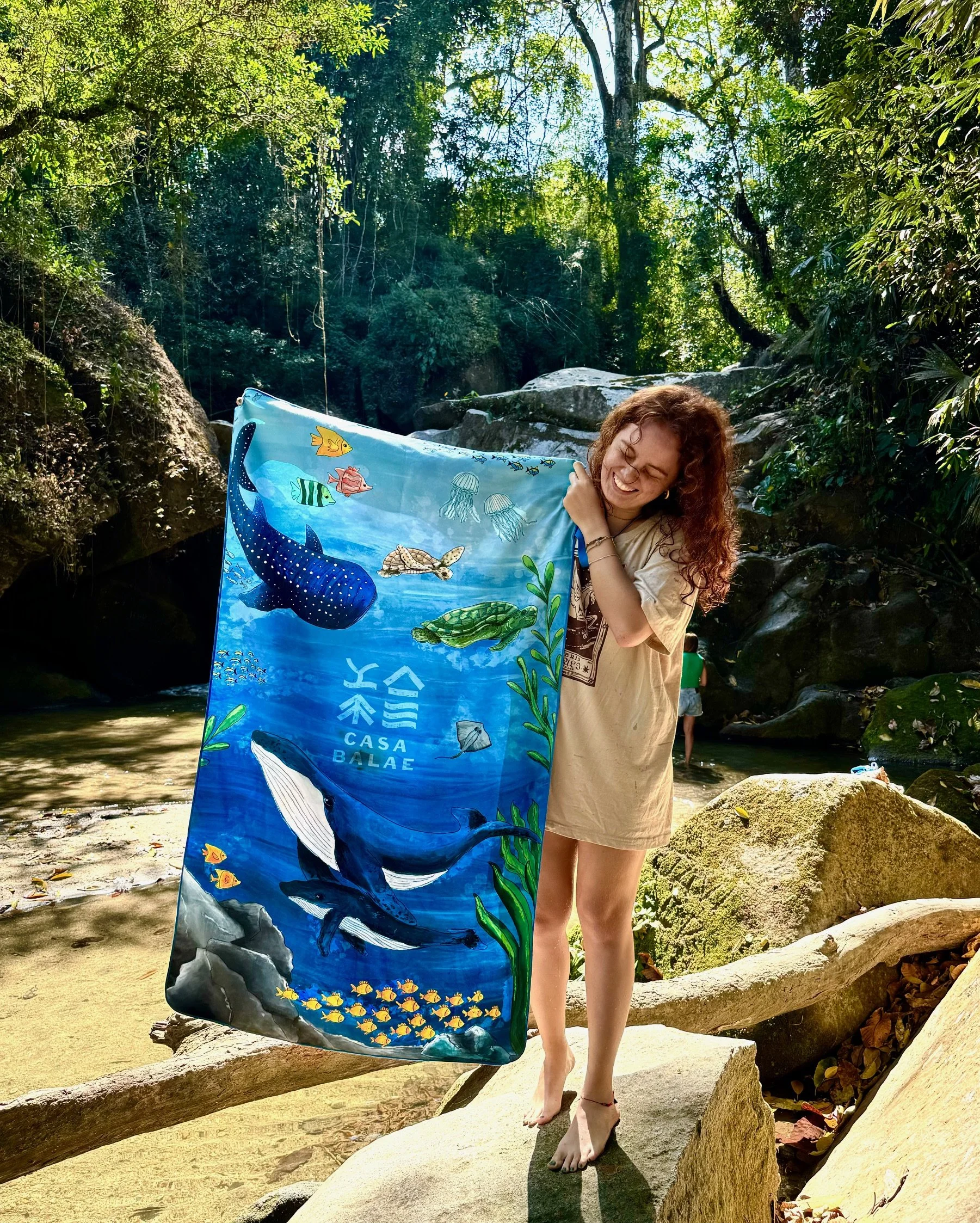 A woman with curly hair smiling holds a large fabric with ocean and whale illustrations in a natural outdoor setting with rocks, trees, and a stream.