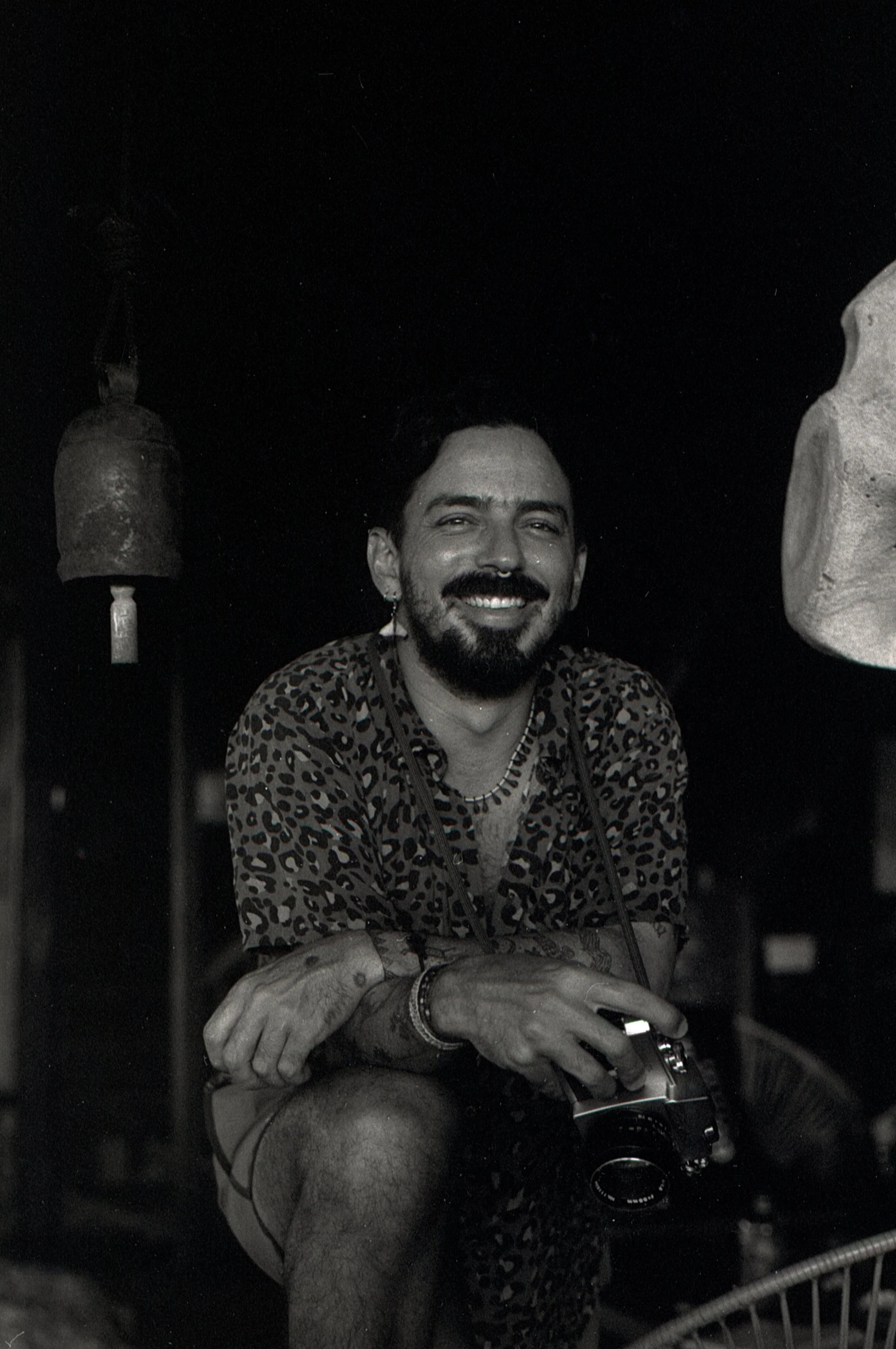 A man with a beard and mustache, wearing a leopard print shirt, smiling and holding a camera, sitting indoors.