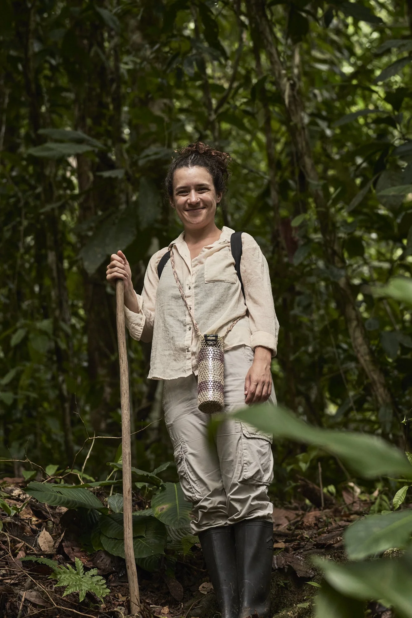 A woman smiling in a jungle, holding a walking stick, wearing a beige long-sleeve shirt, beige cargo pants, and black boots, with a water bottle hanging from her neck and a backpack on her shoulders.