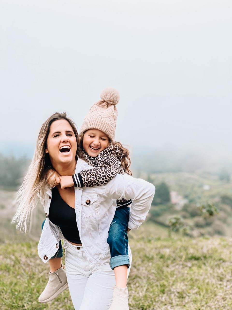 A woman giving a piggyback ride to a young girl in an outdoor grassy area with a misty background of trees and sky. Both are smiling and appear happy.