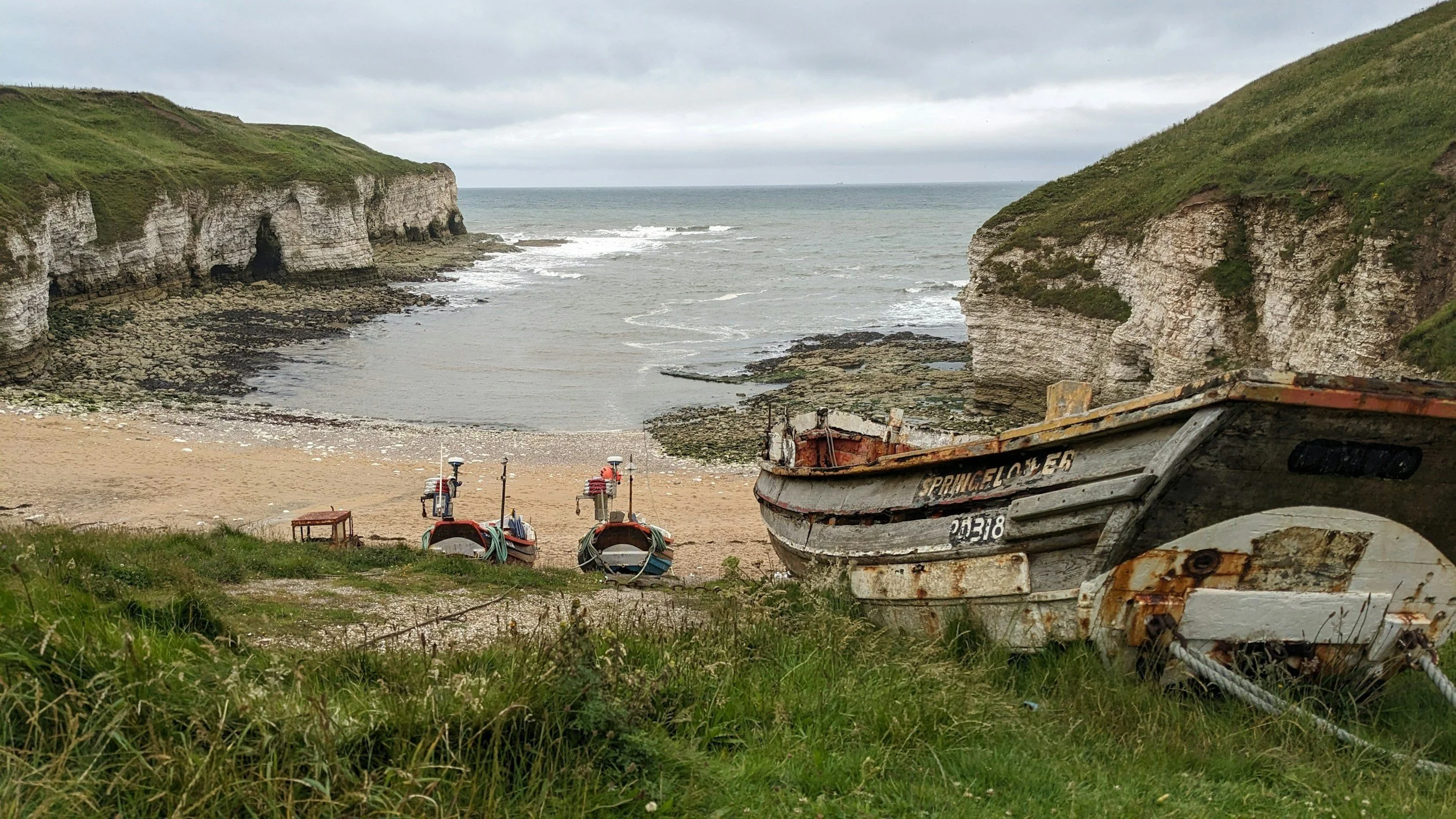 North Landing at Flamborough with boats on grassy foreground at a beach with rocky cliffs and overcast sky in the background.