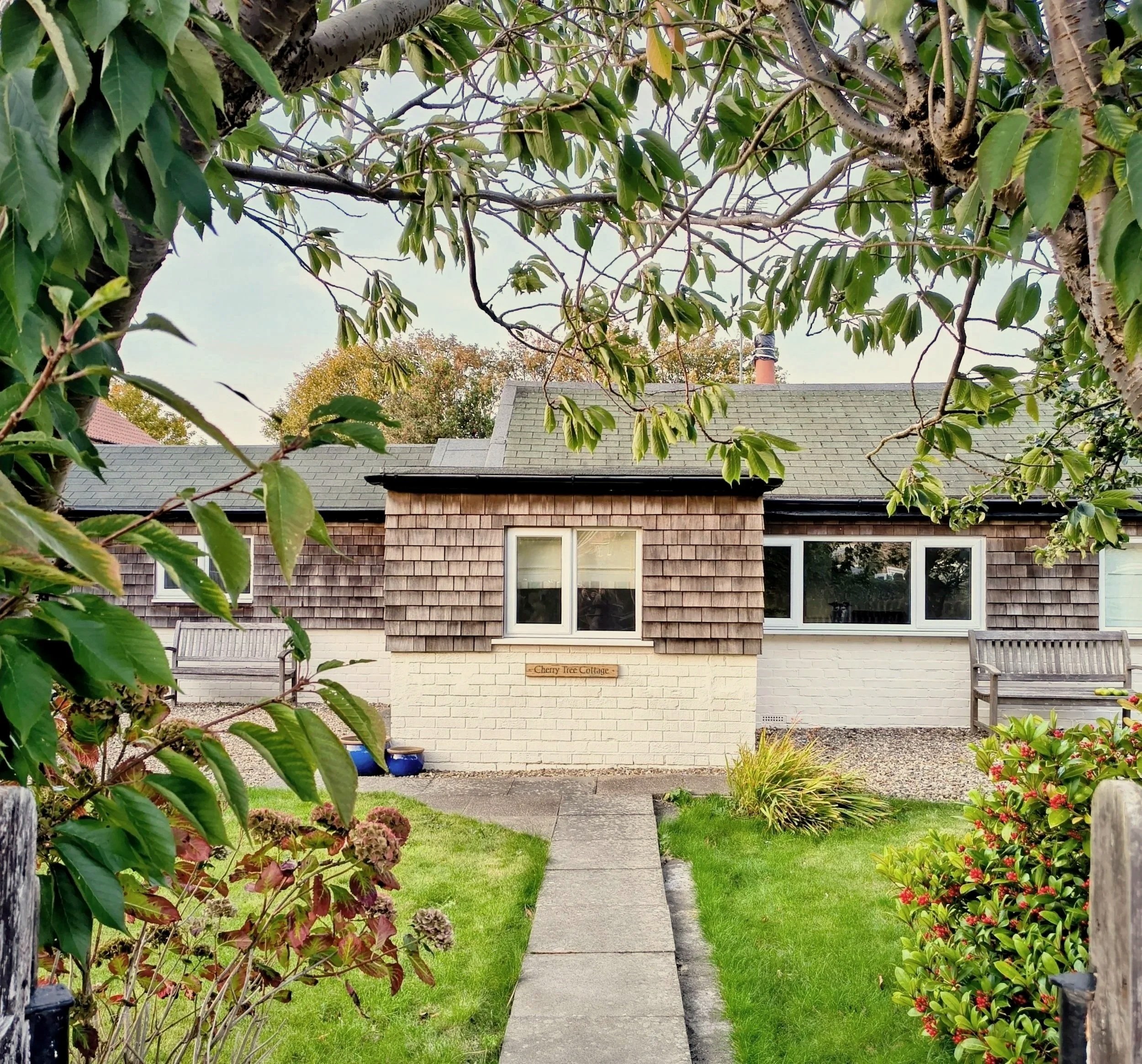Cherry Tree Cottage in Flamborough with wooden shingles on the exterior, and white brick on the lower part. The garden has a gravel area, a green lawn, and outdoor seating including chairs and a bench.