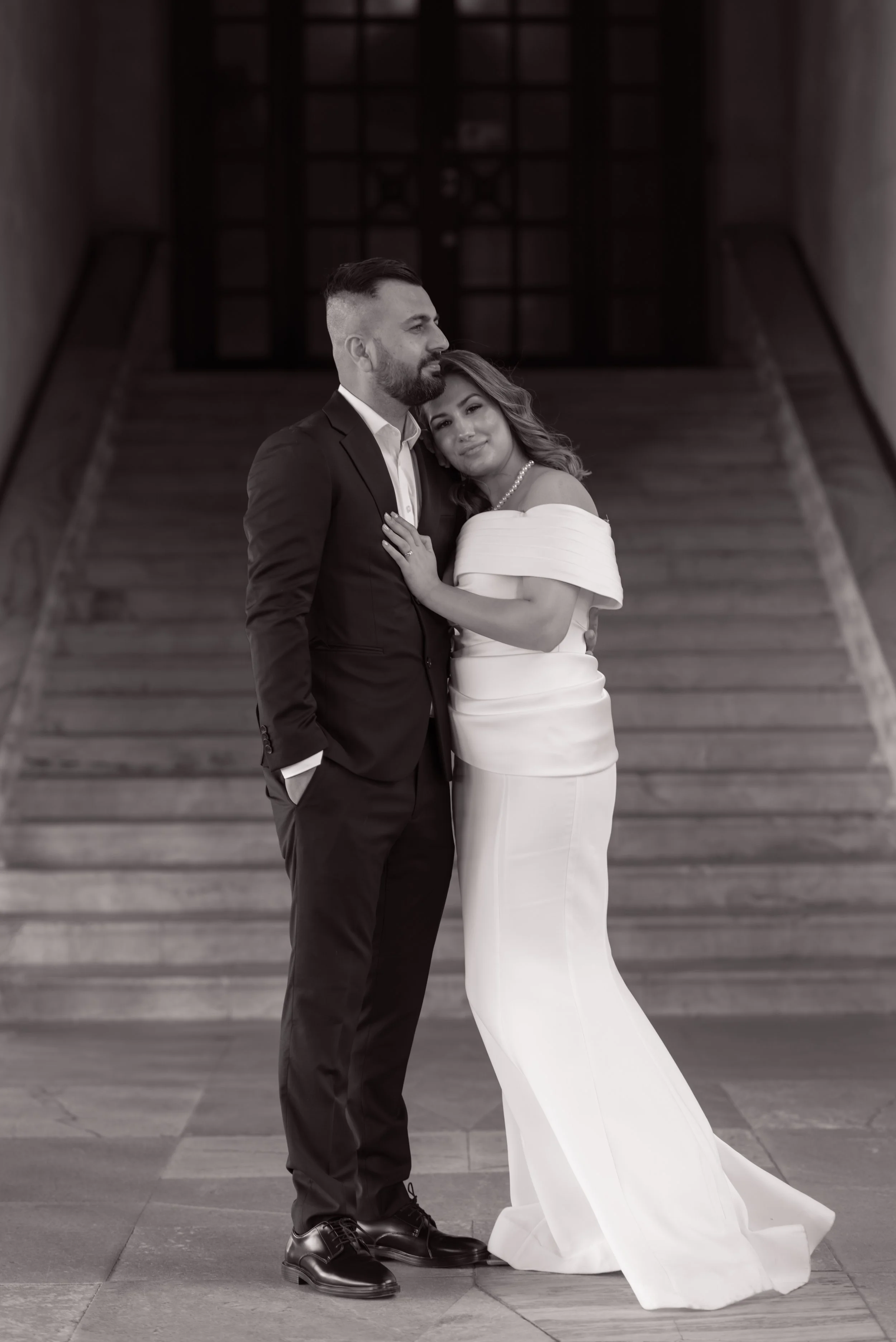 A couple dressed in formal wedding attire standing close together, with the woman resting her head on the man's shoulder, at the top of staircase with wooden railings and glass doors in the background.