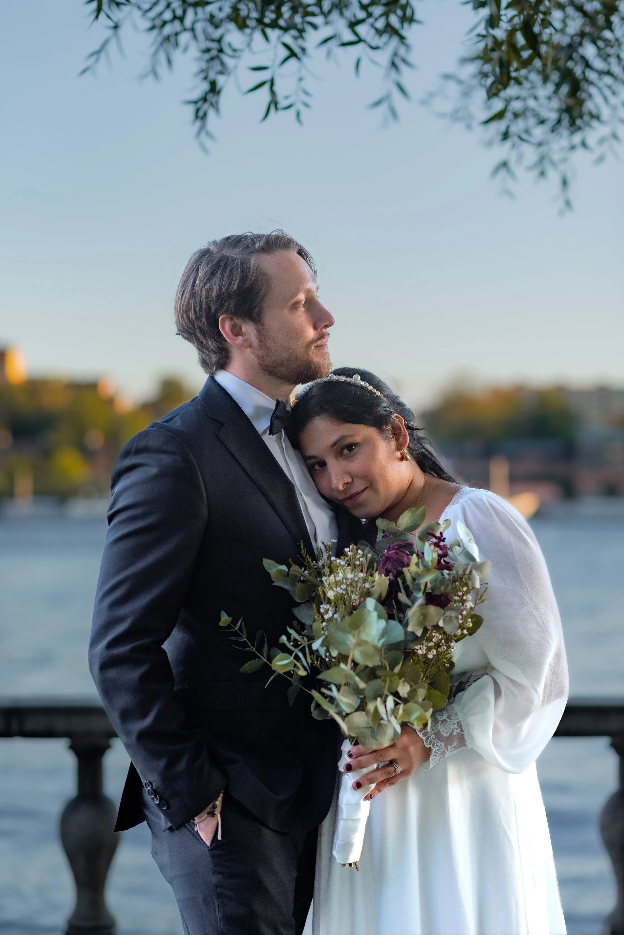 A newlywed couple in wedding attire standing close by a waterfront during sunset. The groom, in a black tuxedo, and the bride, in a white dress, are embracing. The bride holds a bouquet of flowers, and they are framed by overhanging tree branches.