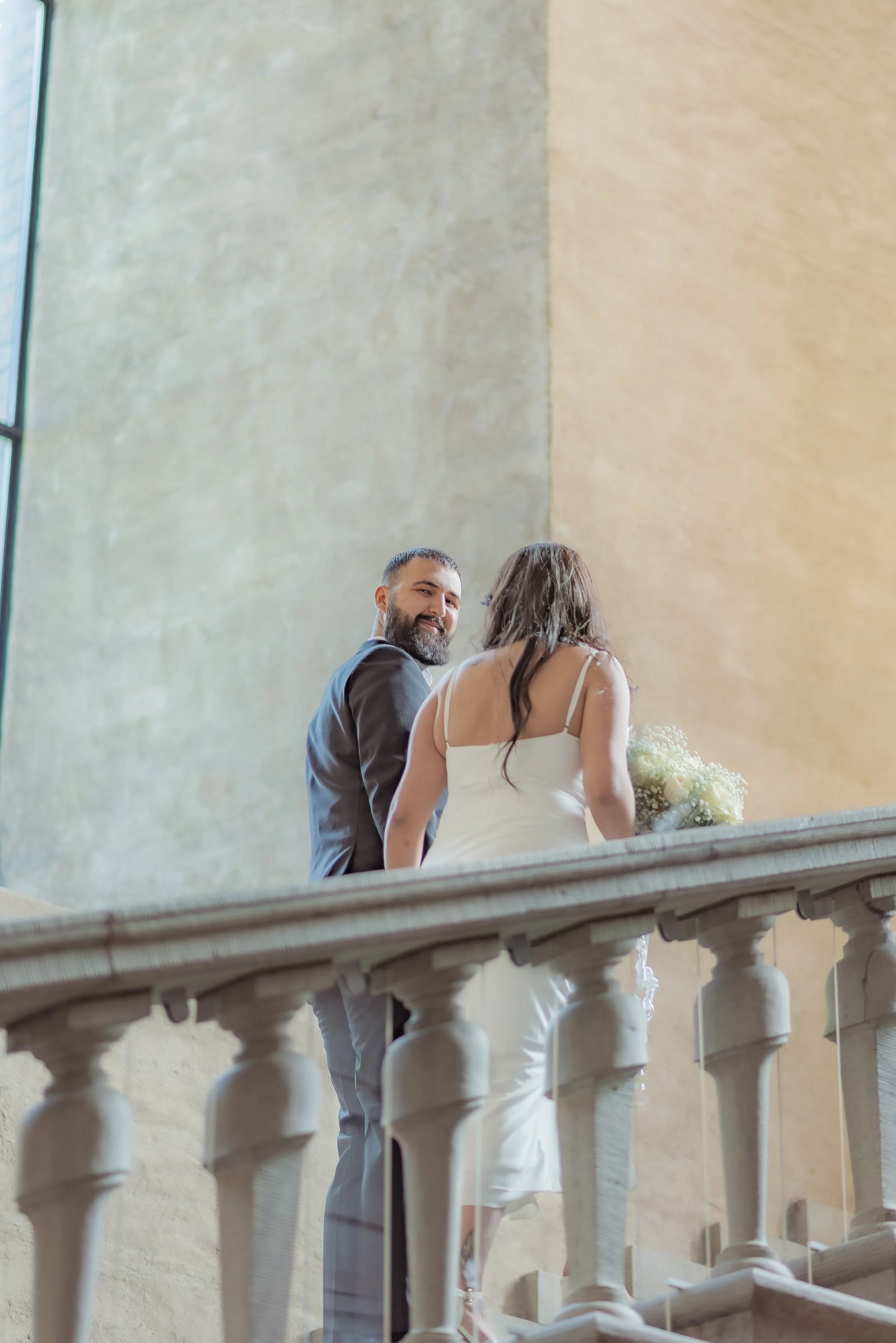 A couple, possibly at a wedding or special event, standing on a balcony with a stone railing inside a building with large windows and beige walls. The man is dressed in a dark suit, smiling at the woman, who is wearing a white dress and holding a bouquet of flowers.