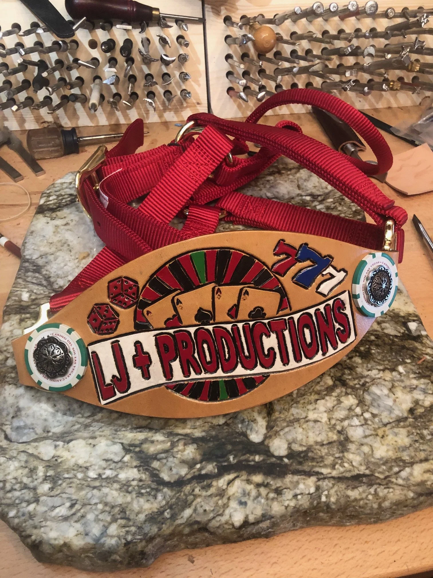 A poker-themedbronc halter with playing cards, dice, and a roulette wheel design, attached to a red harness and displayed on a rock surface. The charm has the text "LJ + PRODUCTIONS" on it.