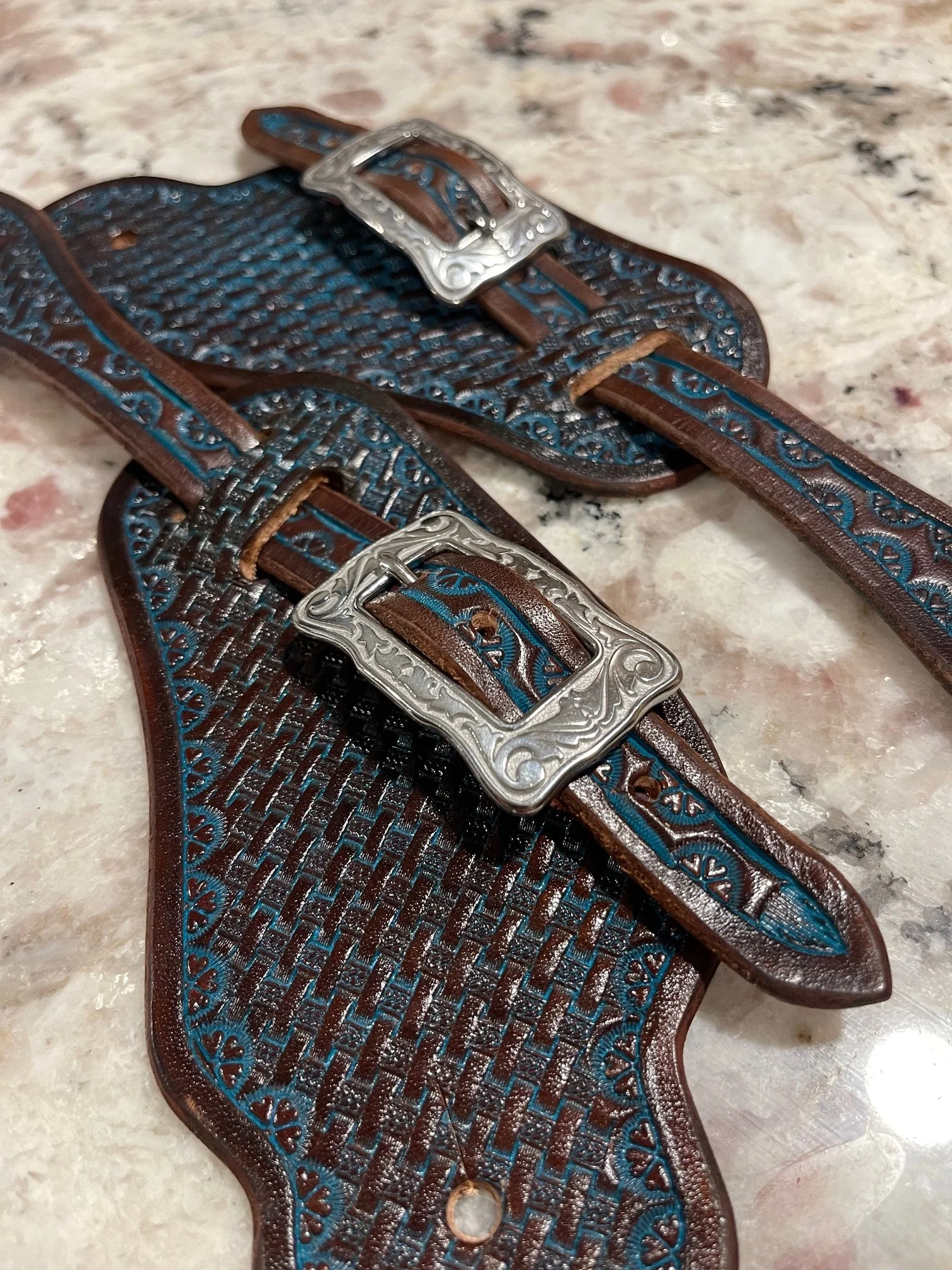 A pair of Western-style leather spur straps with turquoise blue accents and intricate tooling on the straps, lying on a marble surface.