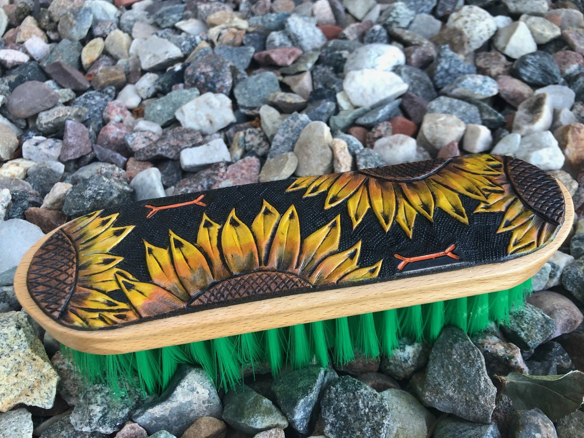 horse brush with a painted sunflower design on the top, green bristles underneath, and a wooden sole, placed on a bed of multicolored rocks.