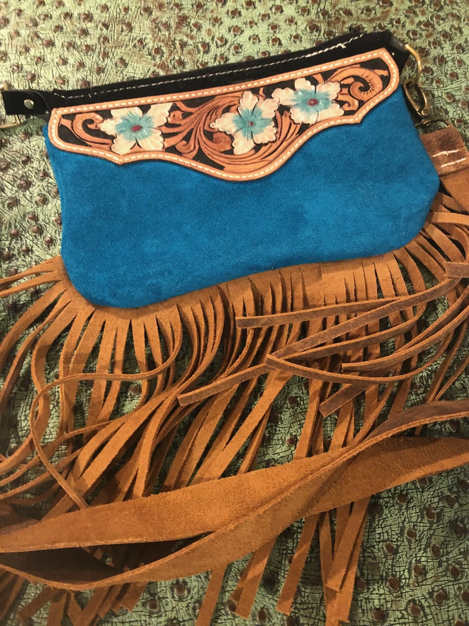 A blue felt handbag with a decorative floral design on the top flap and fringed suede straps resting on a patterned surface.