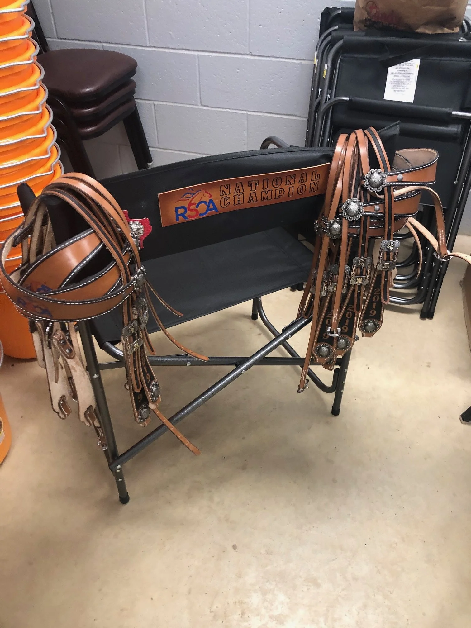 A black bench with a sign that reads 'RSA National Champion' and a collection of brown leather belts with metal conchos hanging over the sides of the bench, situated in a room with stacked chairs and orange trash bins.