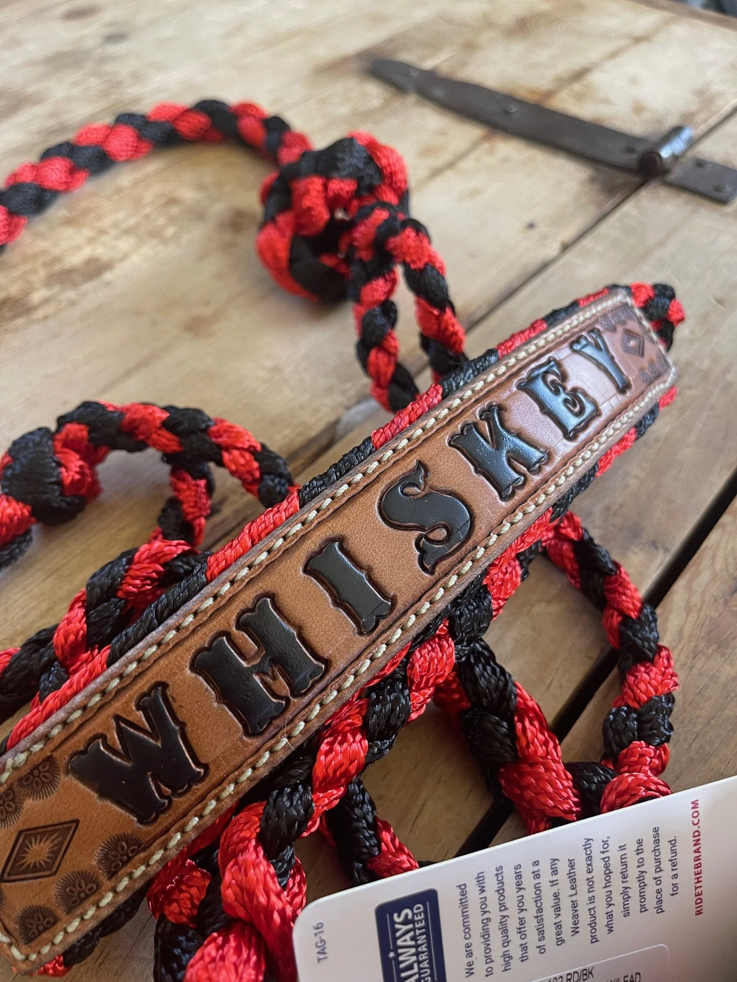 A close-up of a red and black woven rope leash with a leather tag that says "Myester" resting on a wooden surface, with a folding knife in the background.