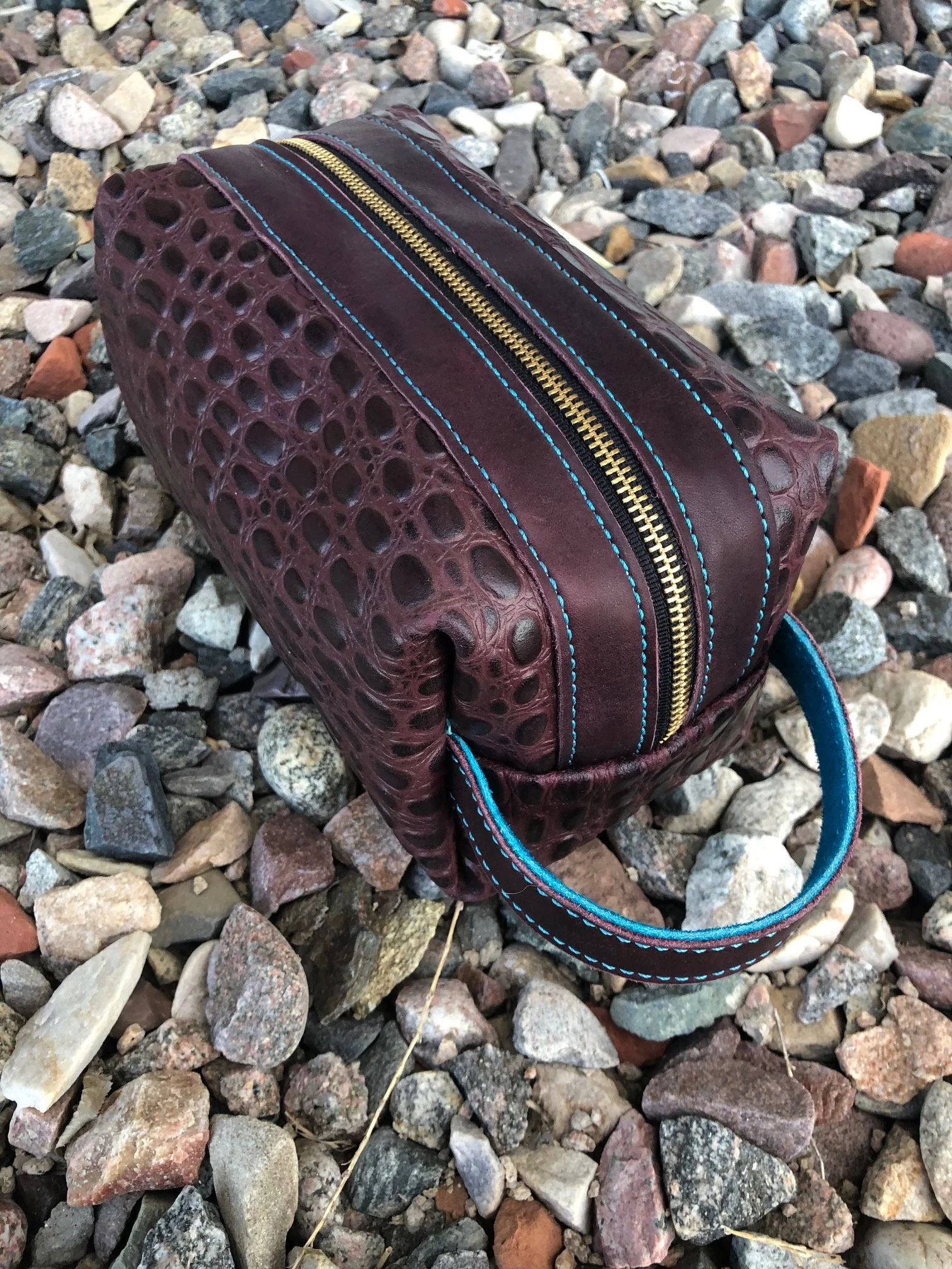 A maroon leather handbag with a crocodile pattern and a blue and black handle, placed on gravel stones outdoors.
