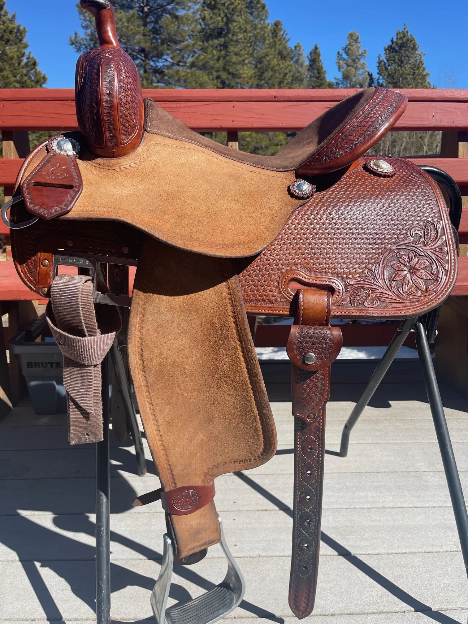 A Western-style leather saddle with detailed carving, placed on a metal stand outdoors with trees and a clear blue sky in the background.