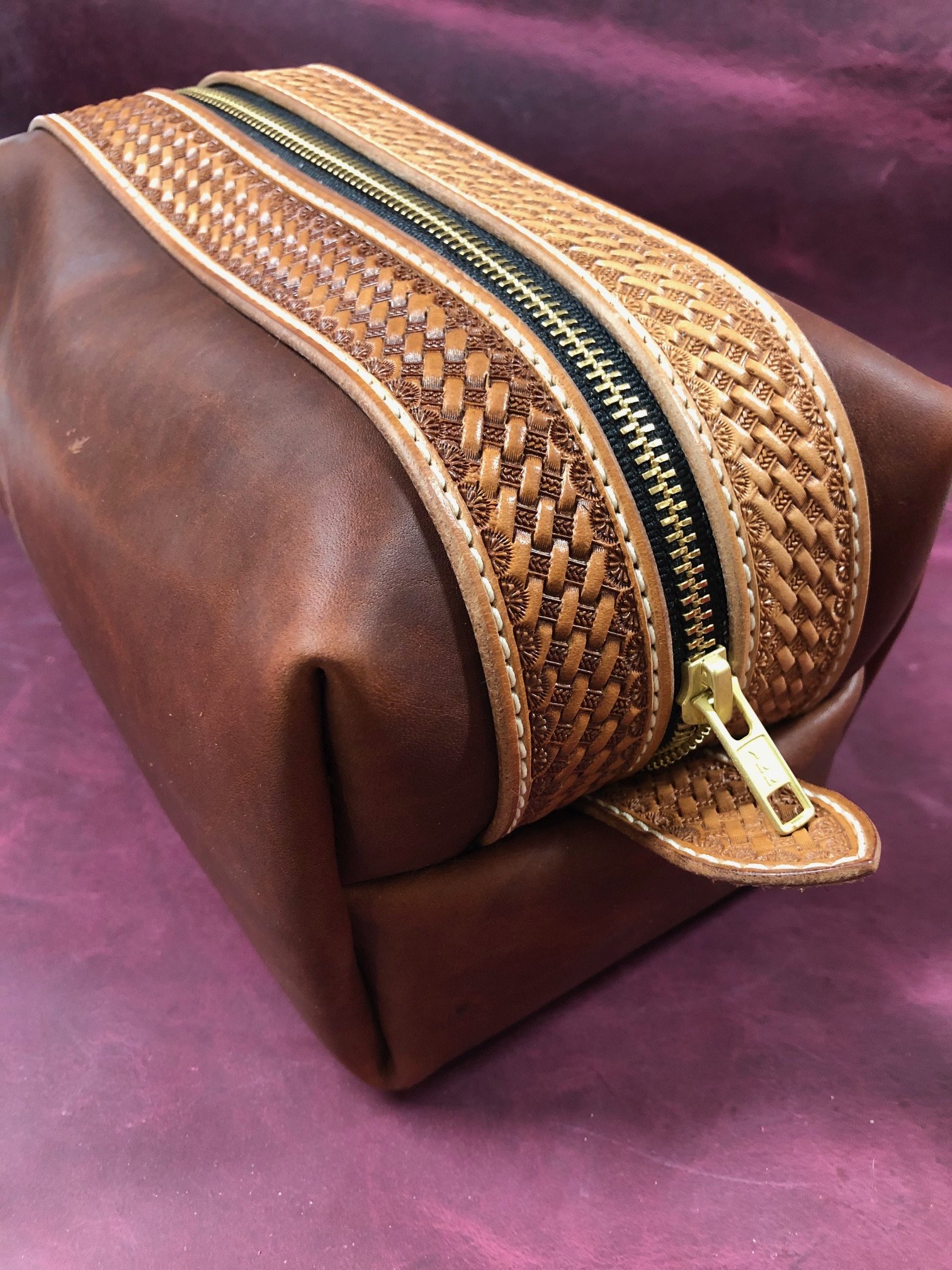 Close-up of a brown leather bag with a decorative woven leather trim and a gold zipper, placed on a purple surface.