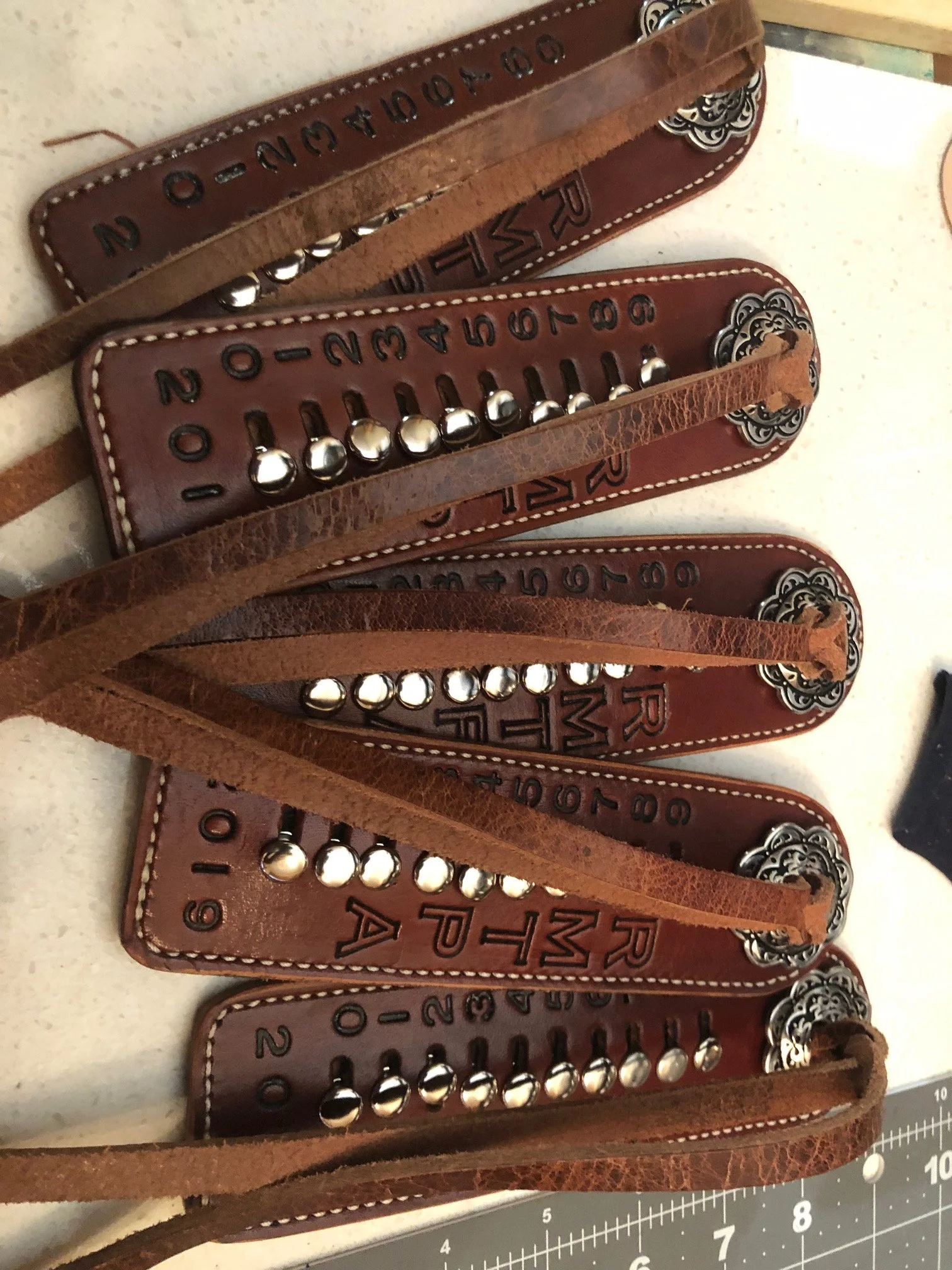 Five leather cow counters, cattle counters each with numbered metal beads and decorative silver accents, placed on a countertop with a measurement ruler visible at the bottom.