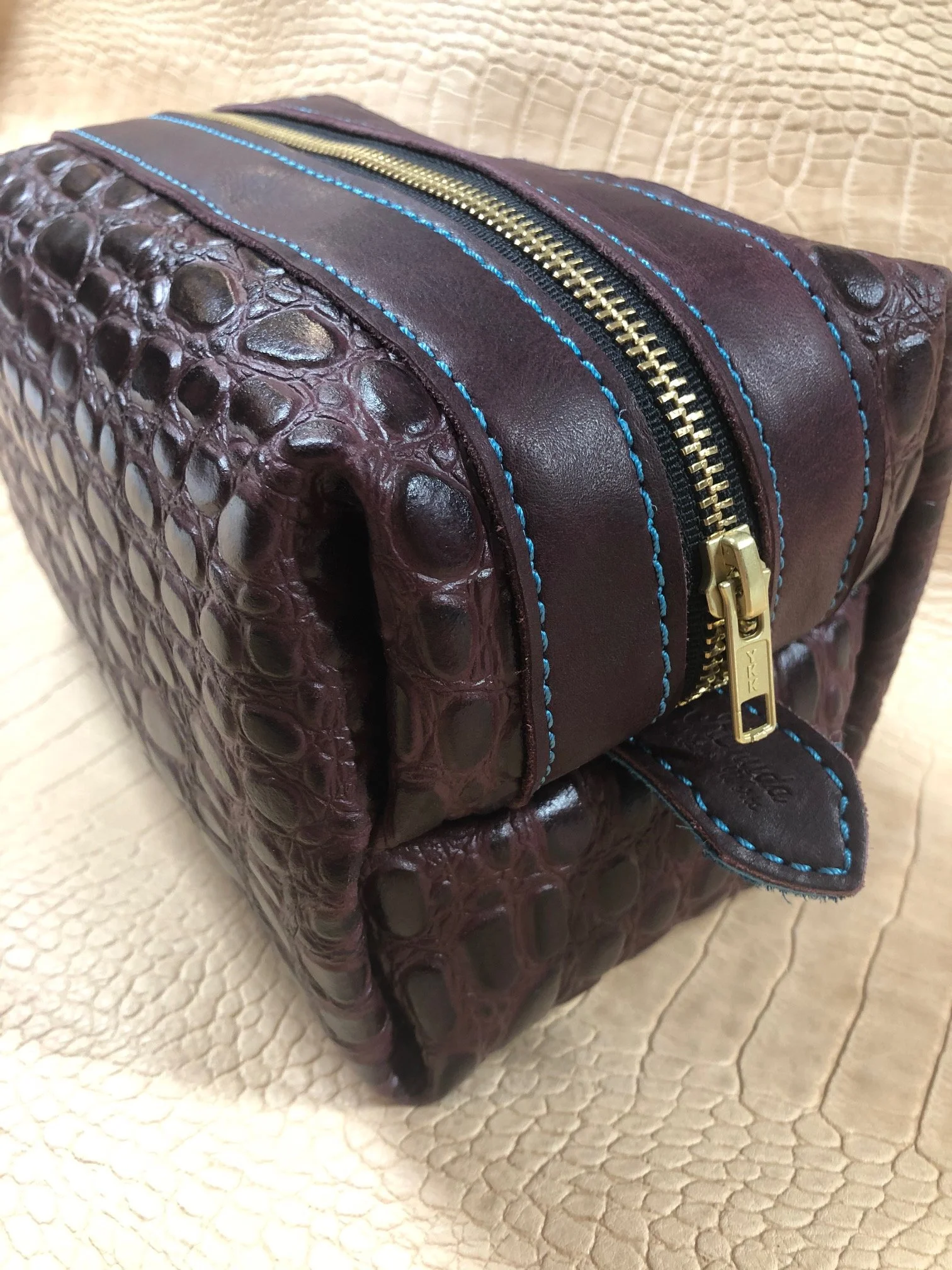 A brown leather bag with an alligator skin texture and blue stitching, featuring a gold zipper.