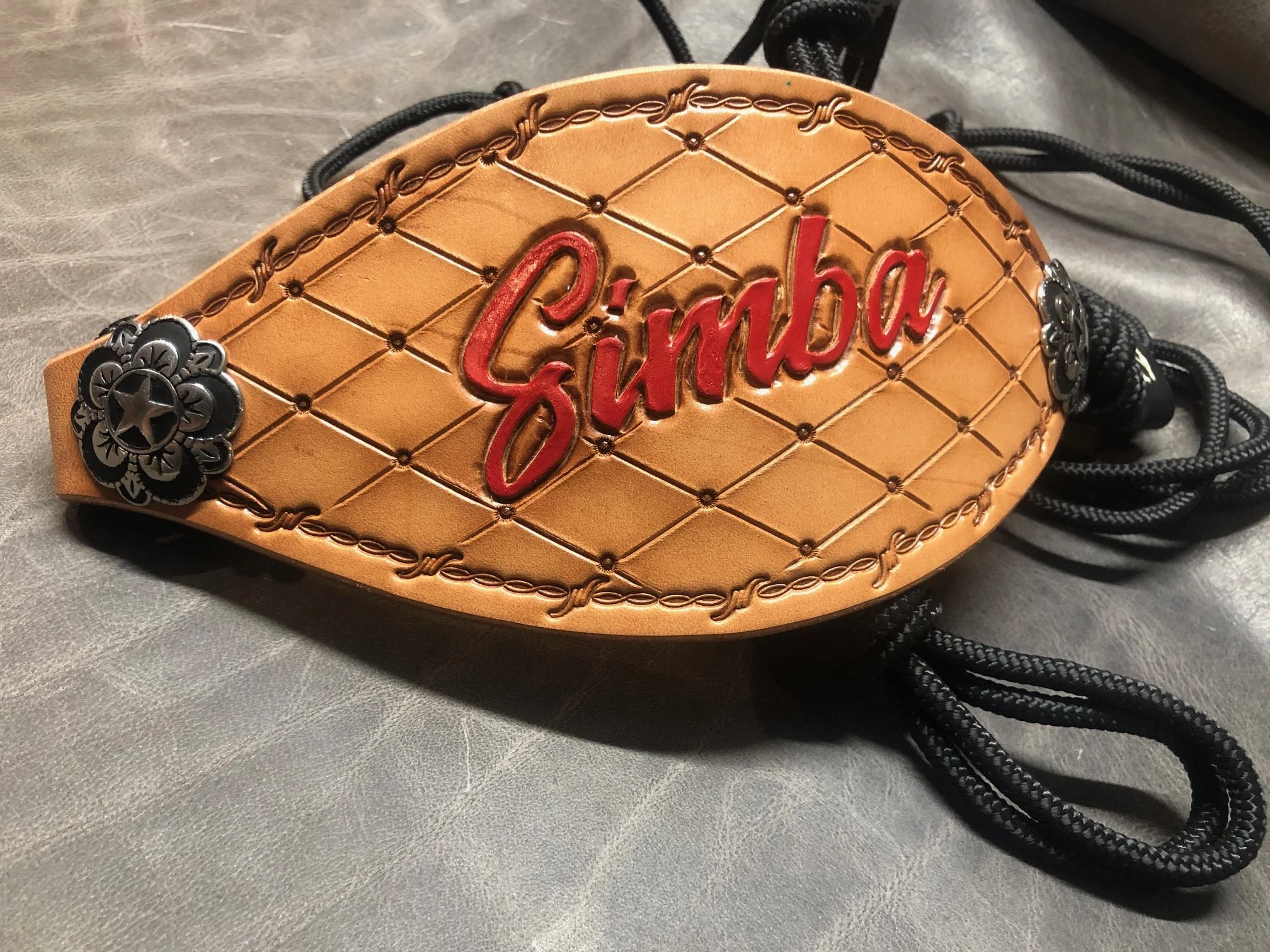 A close-up of a leather bronc halter with the name 'Cinba' embossed in red. The bronc halter is decorated with black and silver conchos and has black drawstrings.