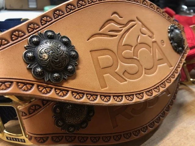 Close-up of a tan leather western-style belt with decorative silver conchos and 'RSA' embossed logo featuring a horse head.