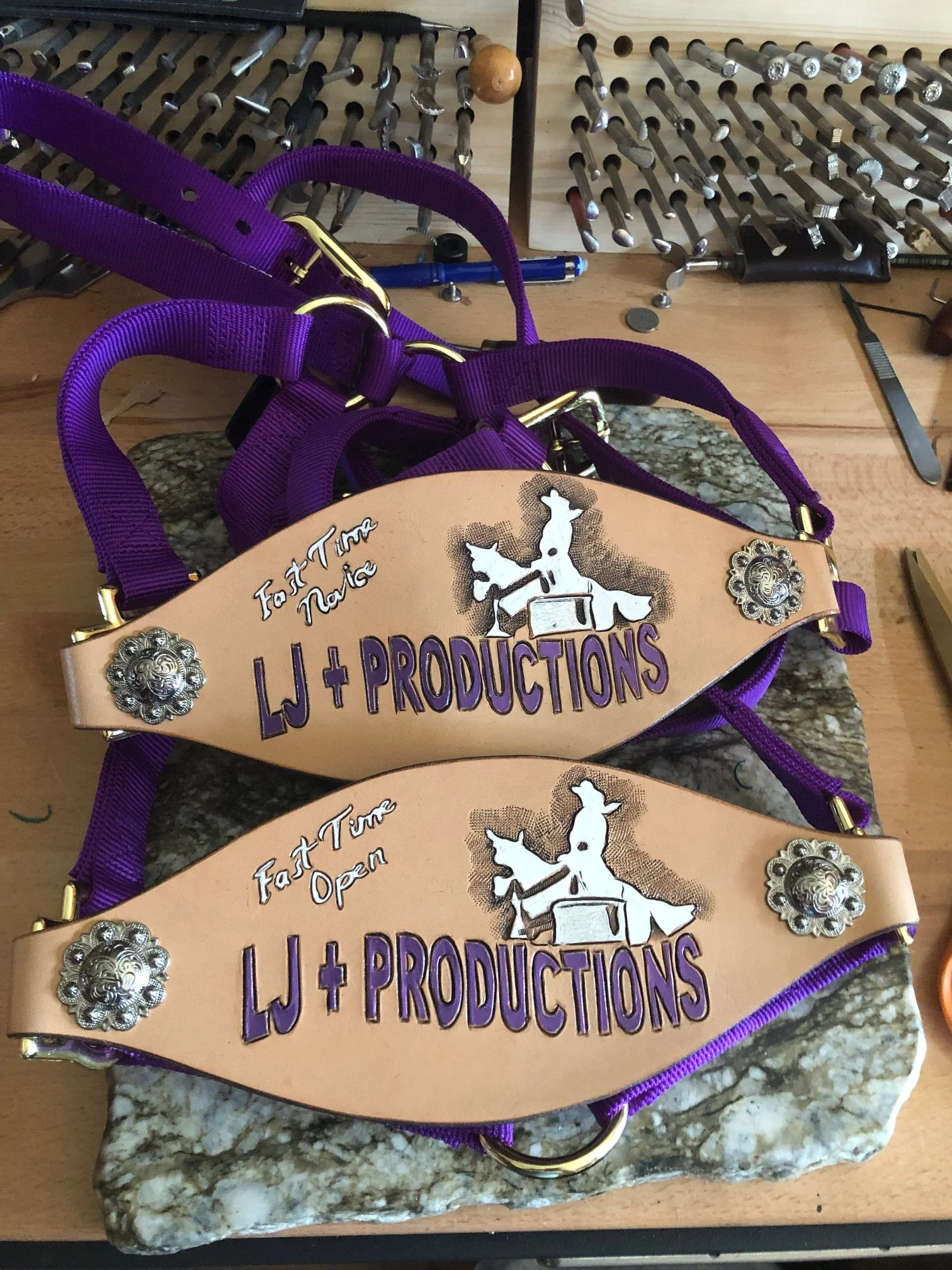 Two handcrafted leather bronc halterswith purple straps, decorative metal accents, and painted leather patches reading 'LJ + PRODUCTIONS' and 'Fast Time' with a silhouette of a dog pulling a sleigh, placed on a stone slab on a workbench.