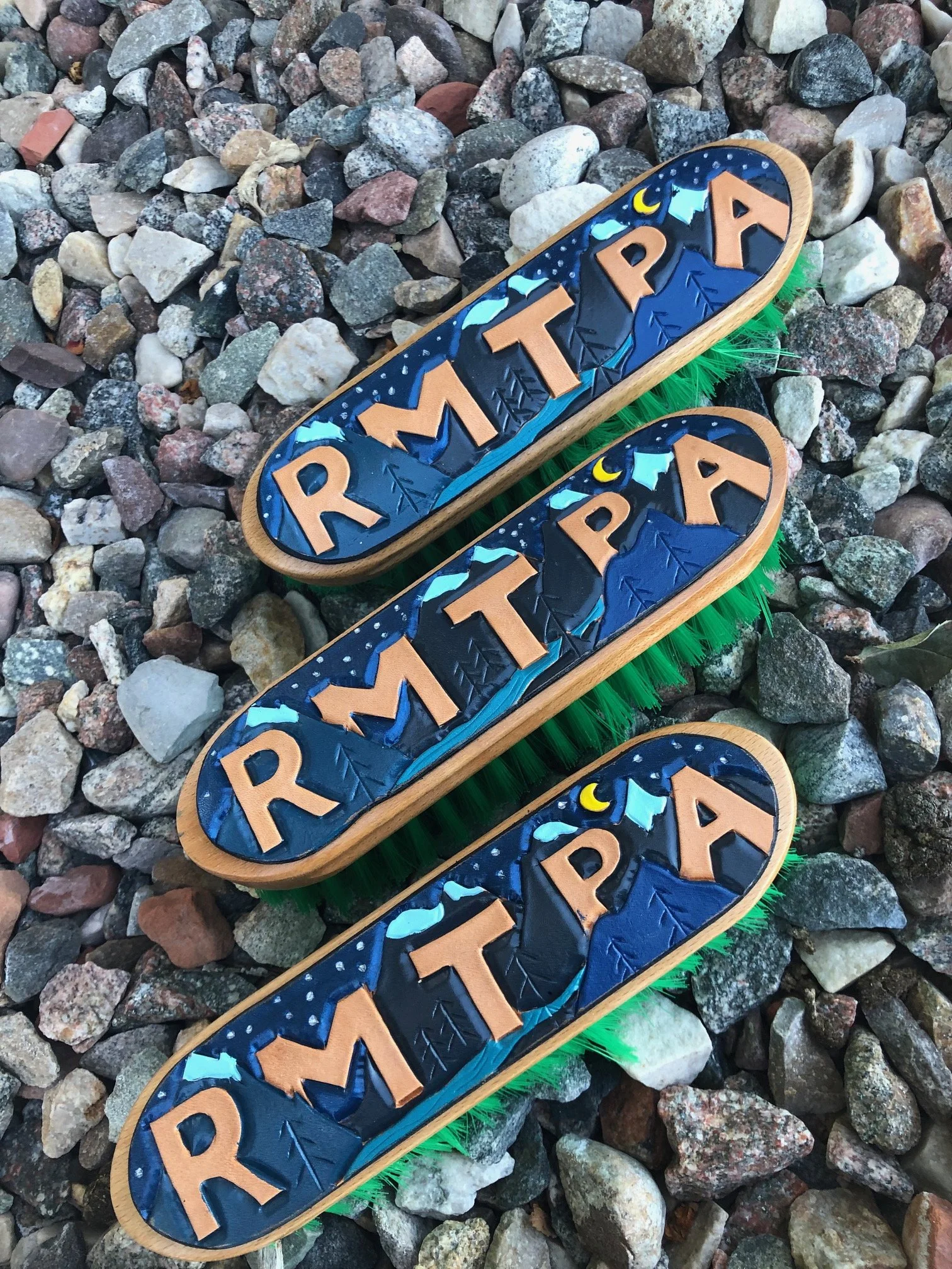 Three horse brushes with the word 'RMTPA' and mountain scenery painted on them, lying on a bed of small rocks.