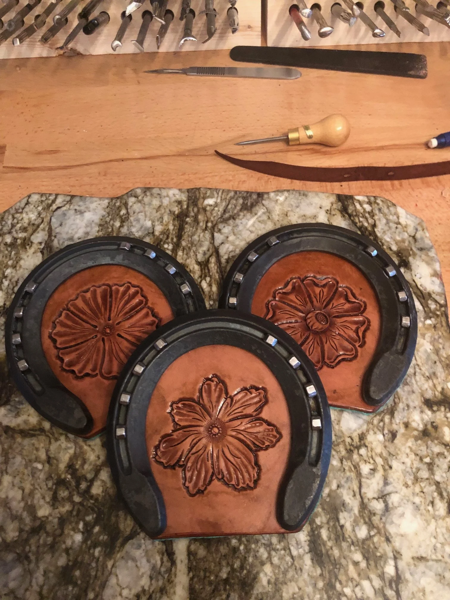 tooled leather horseshoe coasters with floral designs in the center, placed on a marble slab. Various tools, including files and carving tools, are on a wooden workbench in the background.