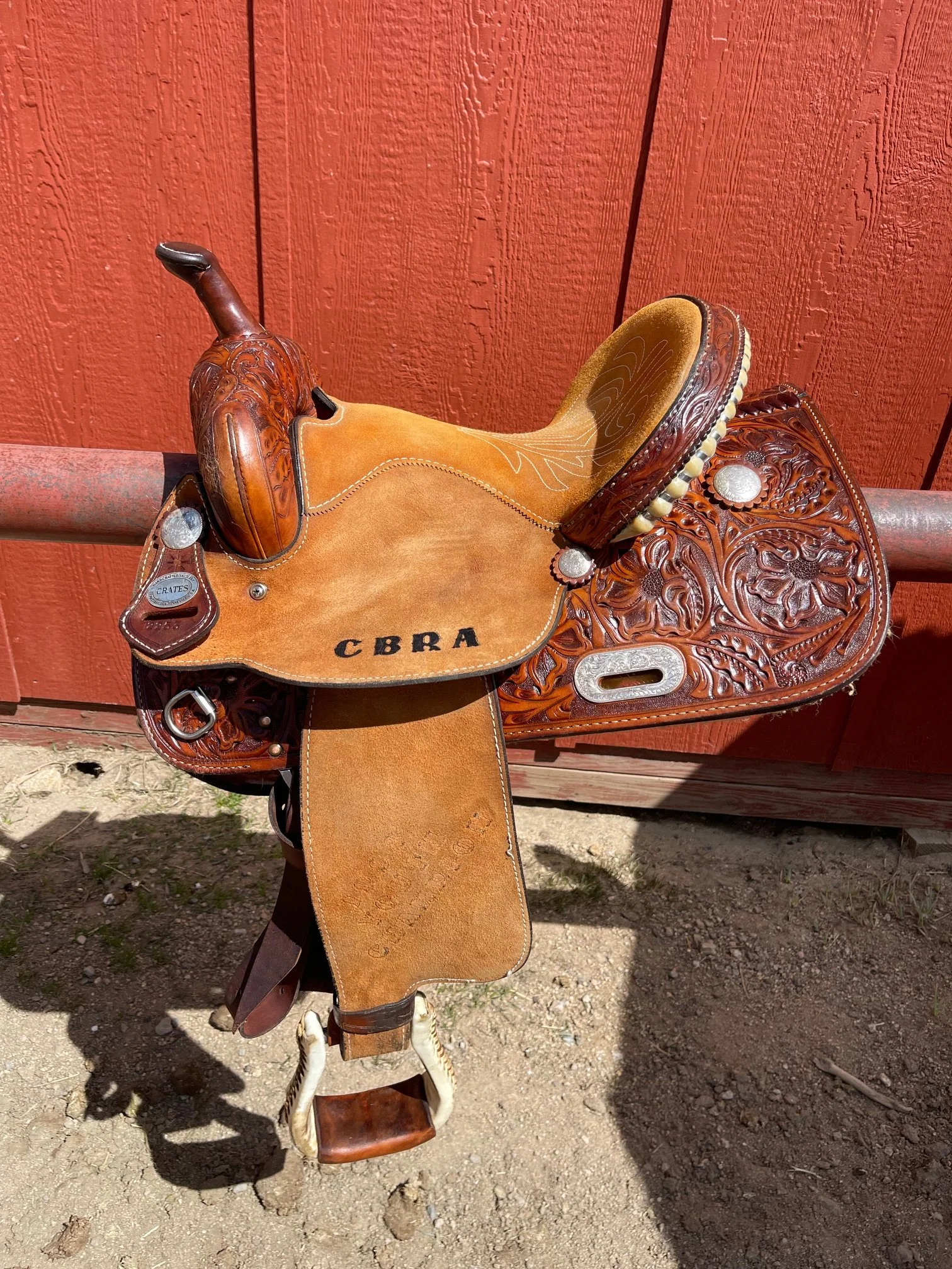 A decorated Western-style horse saddle with intricate leather tooling and a padded seat, resting on a metal rail against a red wooden wall.