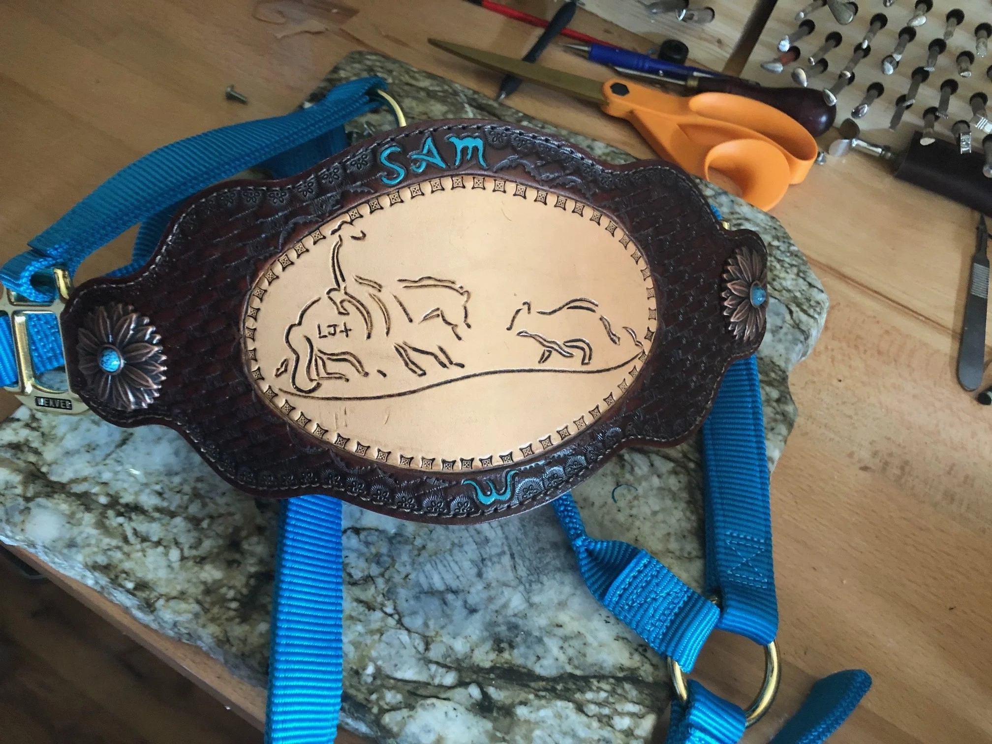 Decorative leather belt buckle with engraved cattle and cow scene, featuring the name 'SAM' in blue at the top, placed on a workbench with tools in the background.