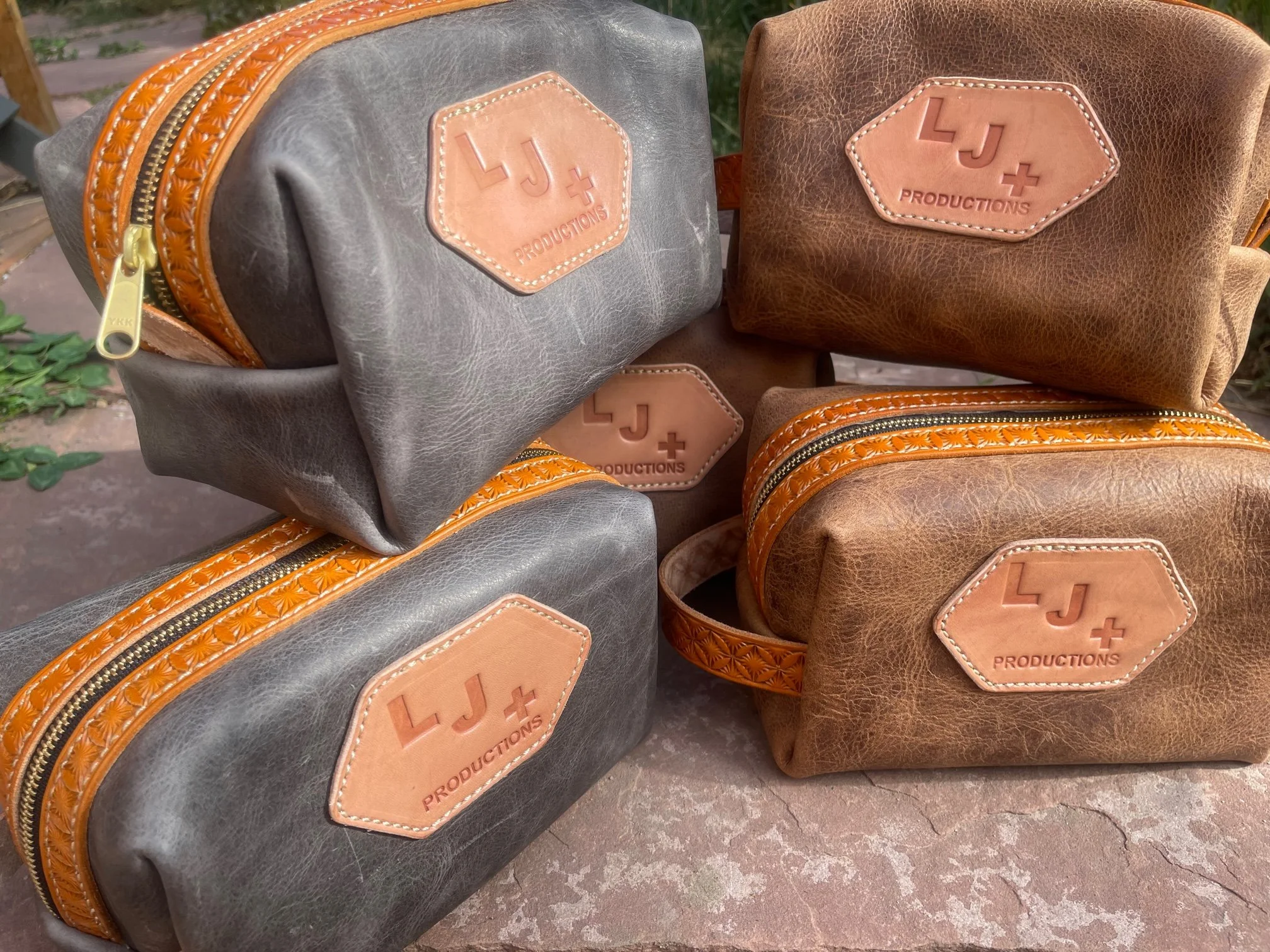 Four leather bags in gray and brown colors with orange accents and a leather patch labeled 'LJ+ PRODUCTIONS' arranged outdoors on a stone surface.
