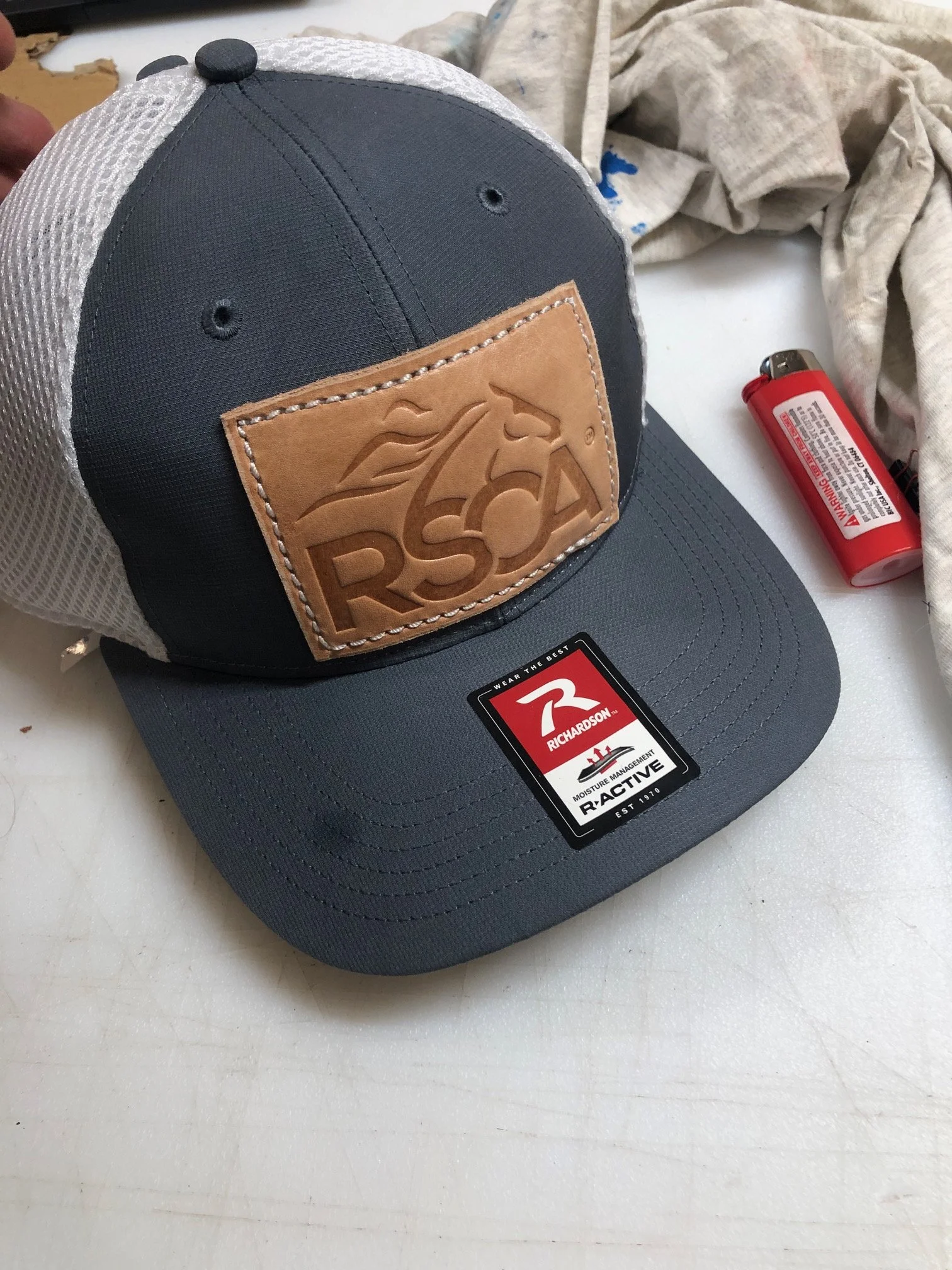 A gray and white Richardson cap with a leather patch featuring a horse and RSD logo, on a white surface next to a red lighter and a wrinkled cloth.
