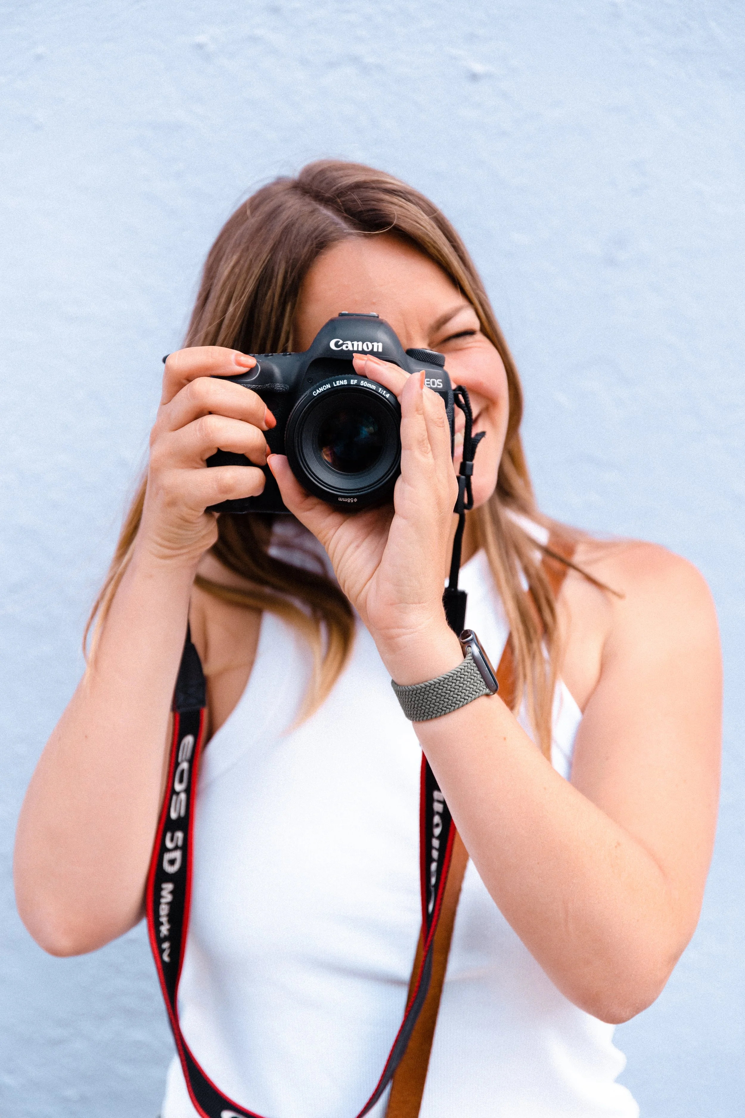 Une femme souriante qui tient un appareil photo Canon devant son visage, portant un t-shirt blanc et un bracelet gris, avec un fond bleu clair.