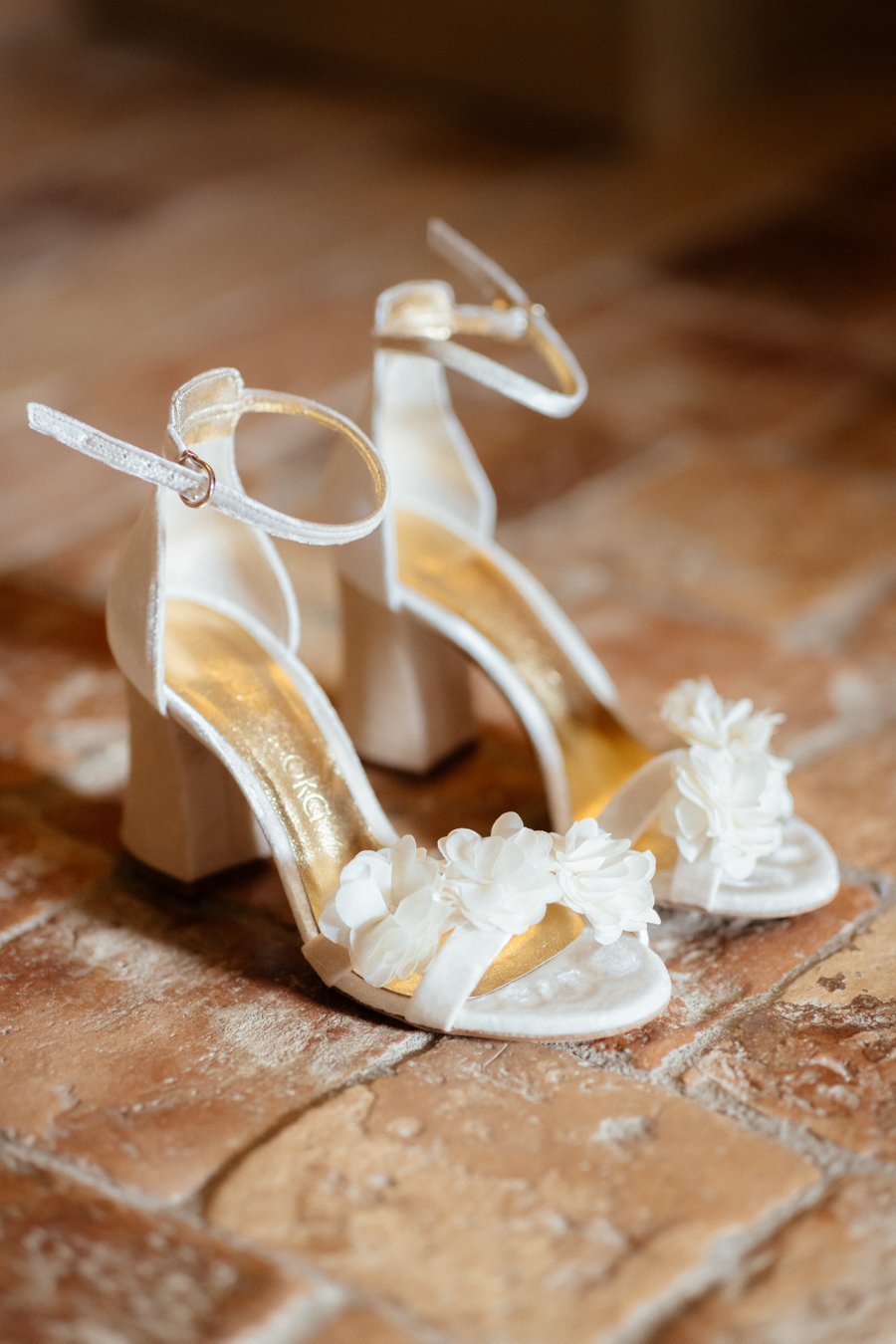Une paire de sandales de mariage blanches avec des fleurs decoratives, posée sur un sol en briques.