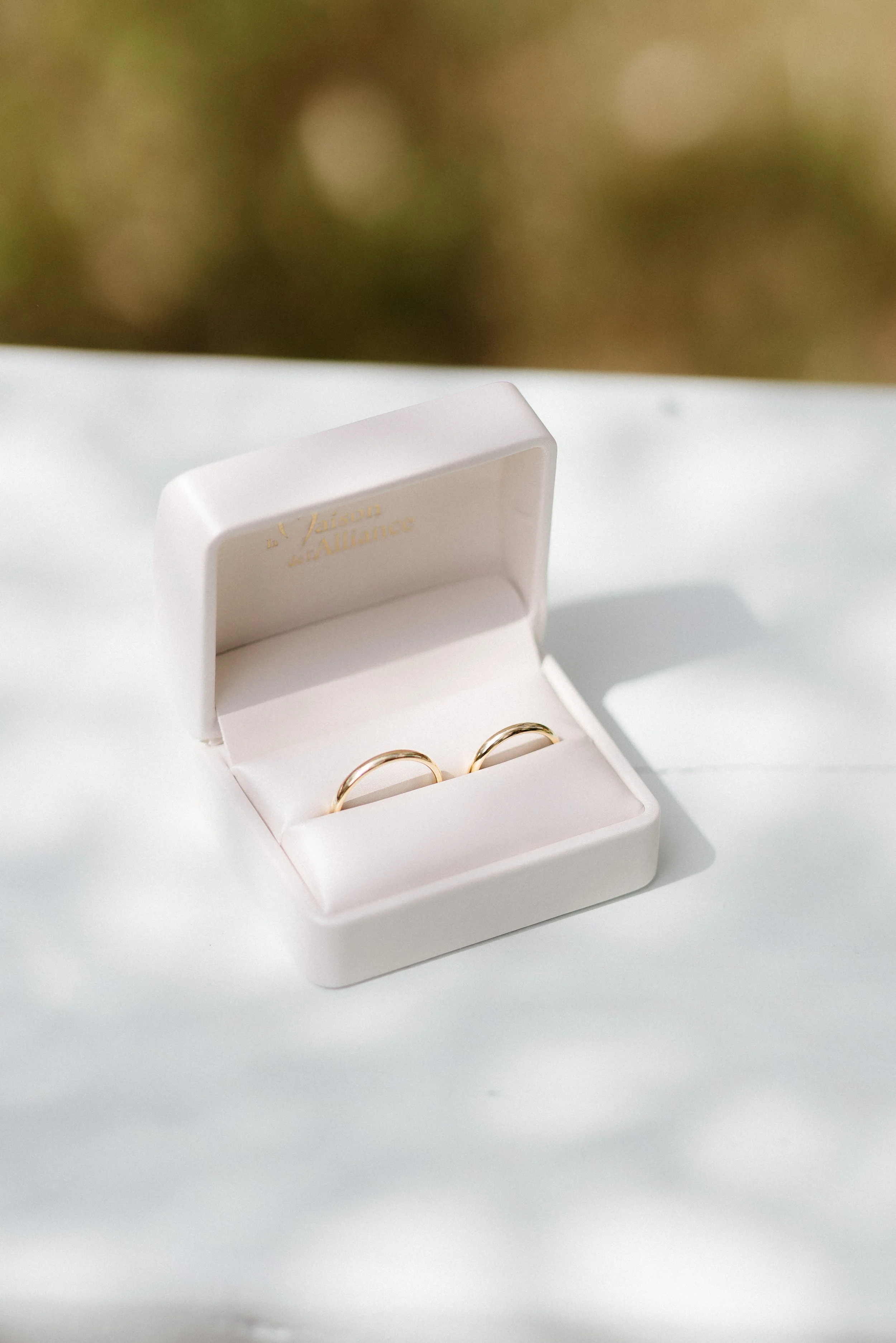 Une boîte à bijoux blanche contenant deux anneaux dorés, posée sur une surface blanche, avec un fond flou de verdure.
