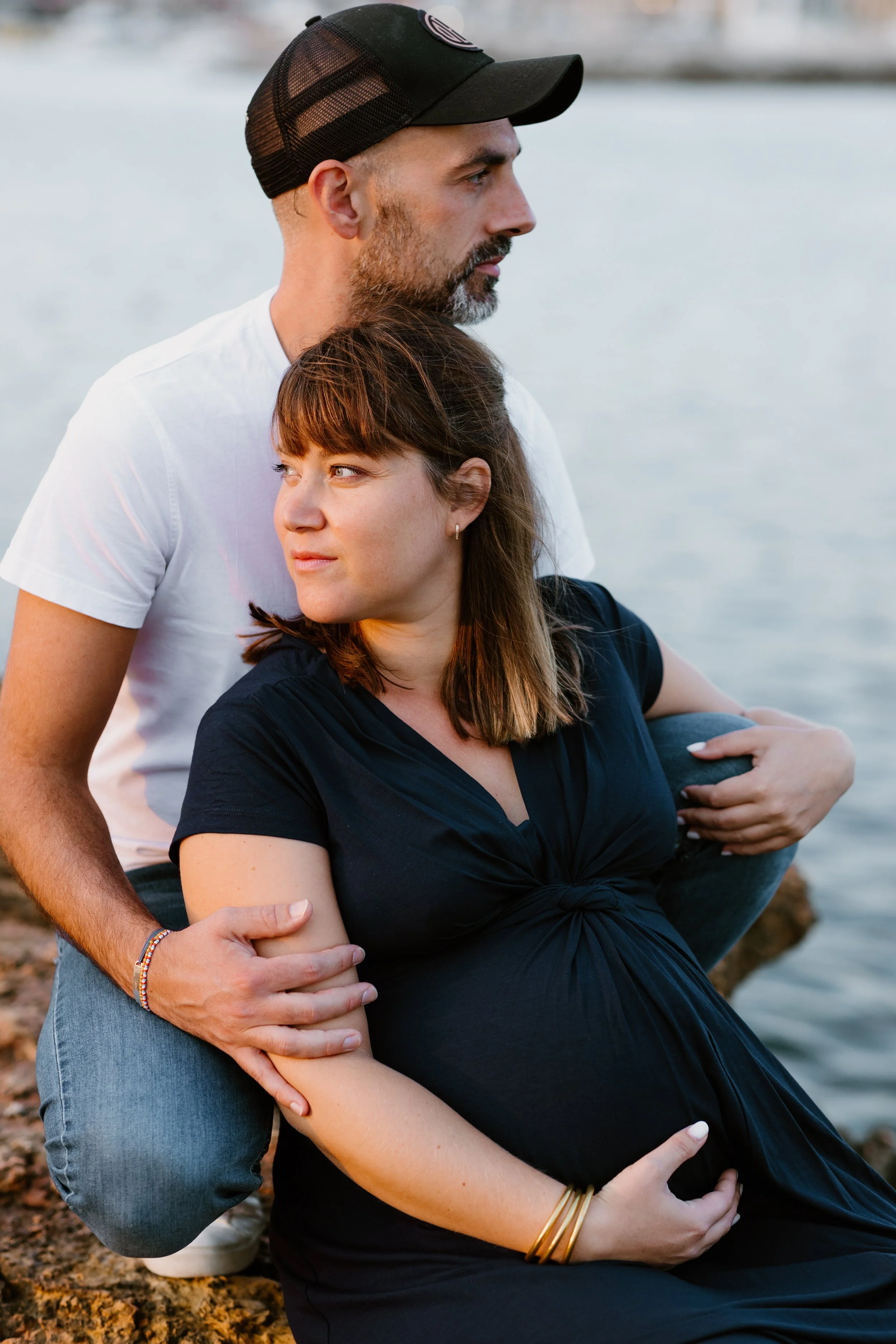 Un couple regarde au loin près d'un lac, la femme est enceinte et porte un vêtement noir, l'homme porte un t-shirt blanc et un chapeau noir. Ils sont assis sur des rochers au bord de l'eau.