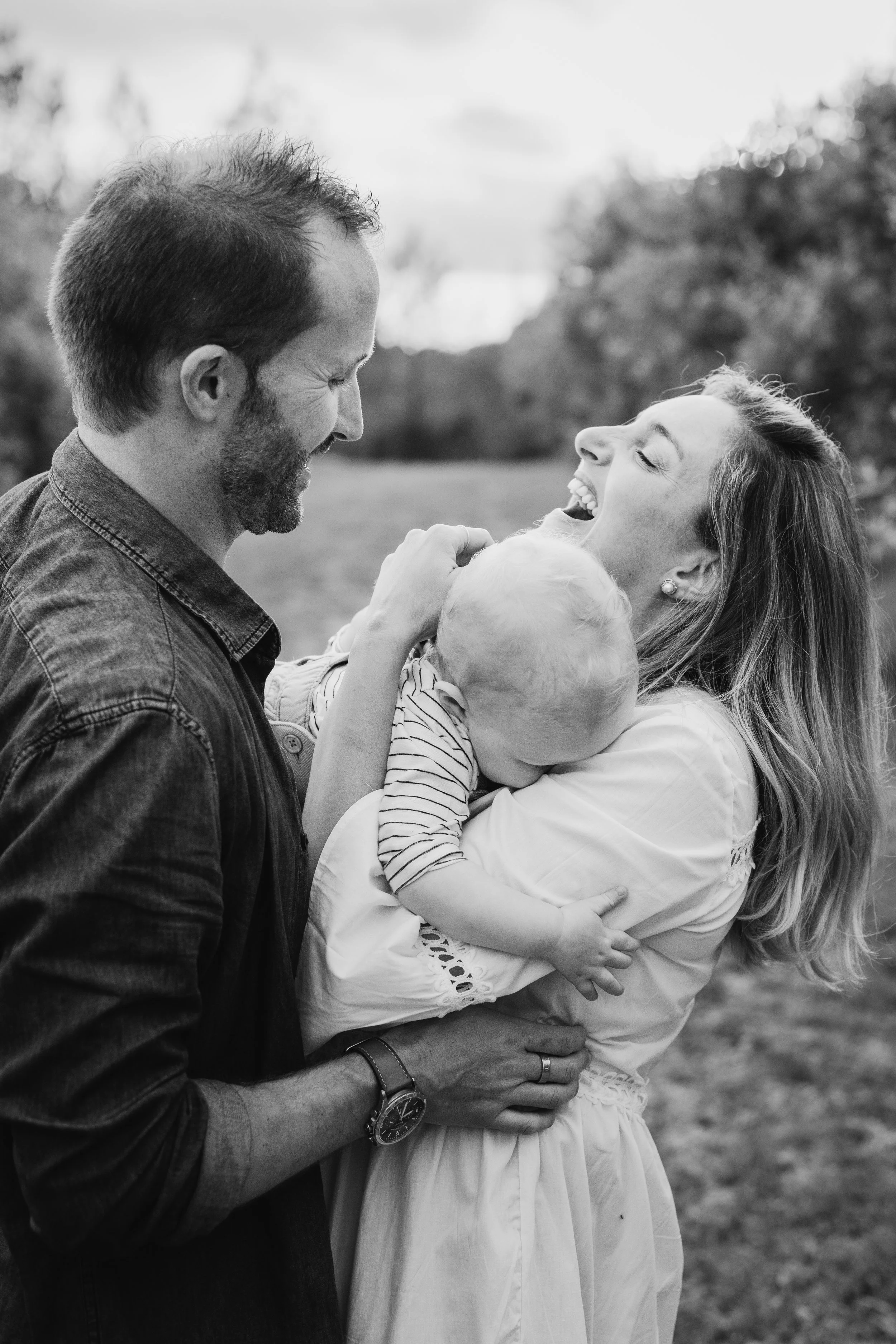 Un couple souriant et riant, tenant un bébé, dans un parc en plein air.