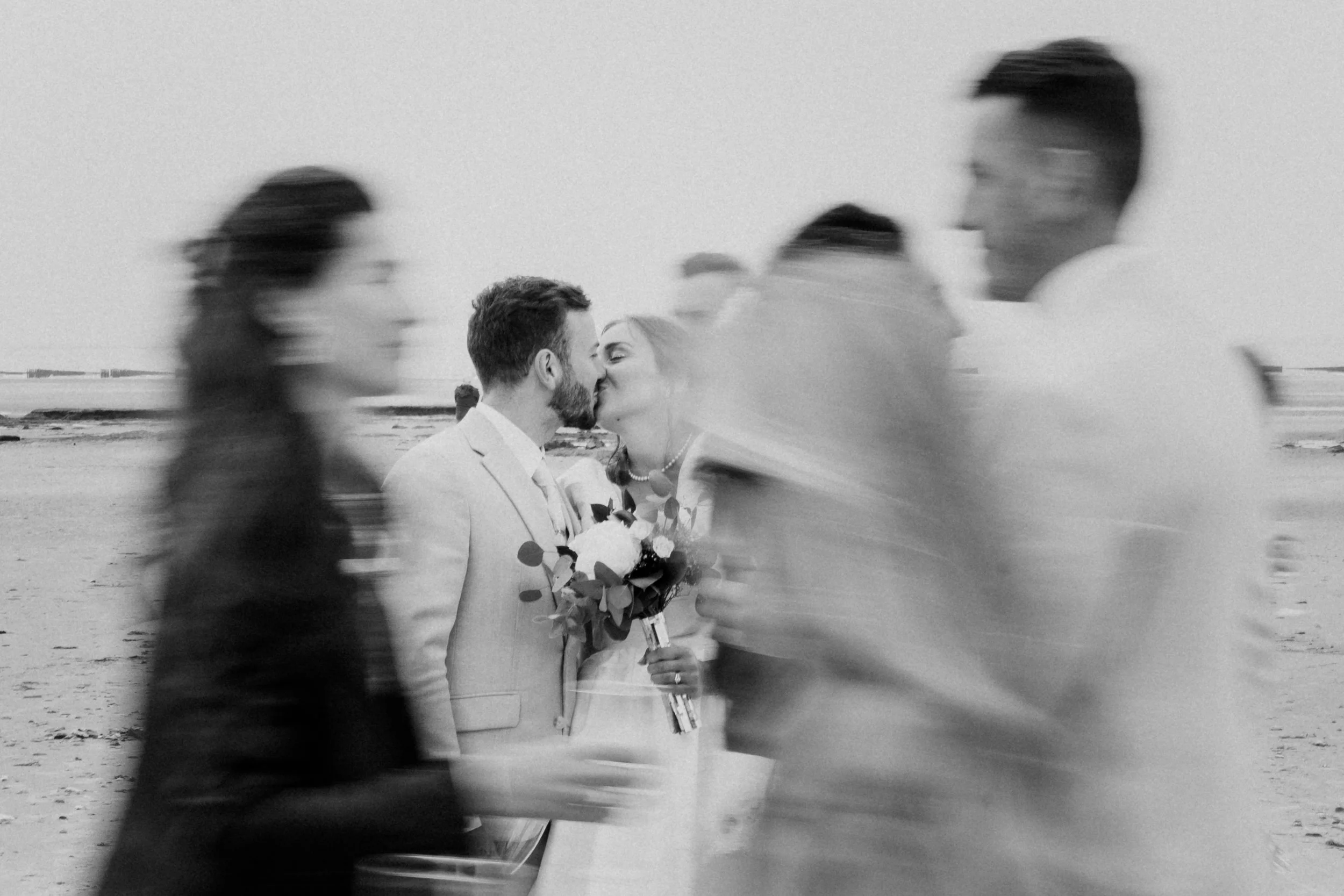 Un couple de mariés s'embrasse sur une plage, entouré de plusieurs personnes floues qui passent devant lui.