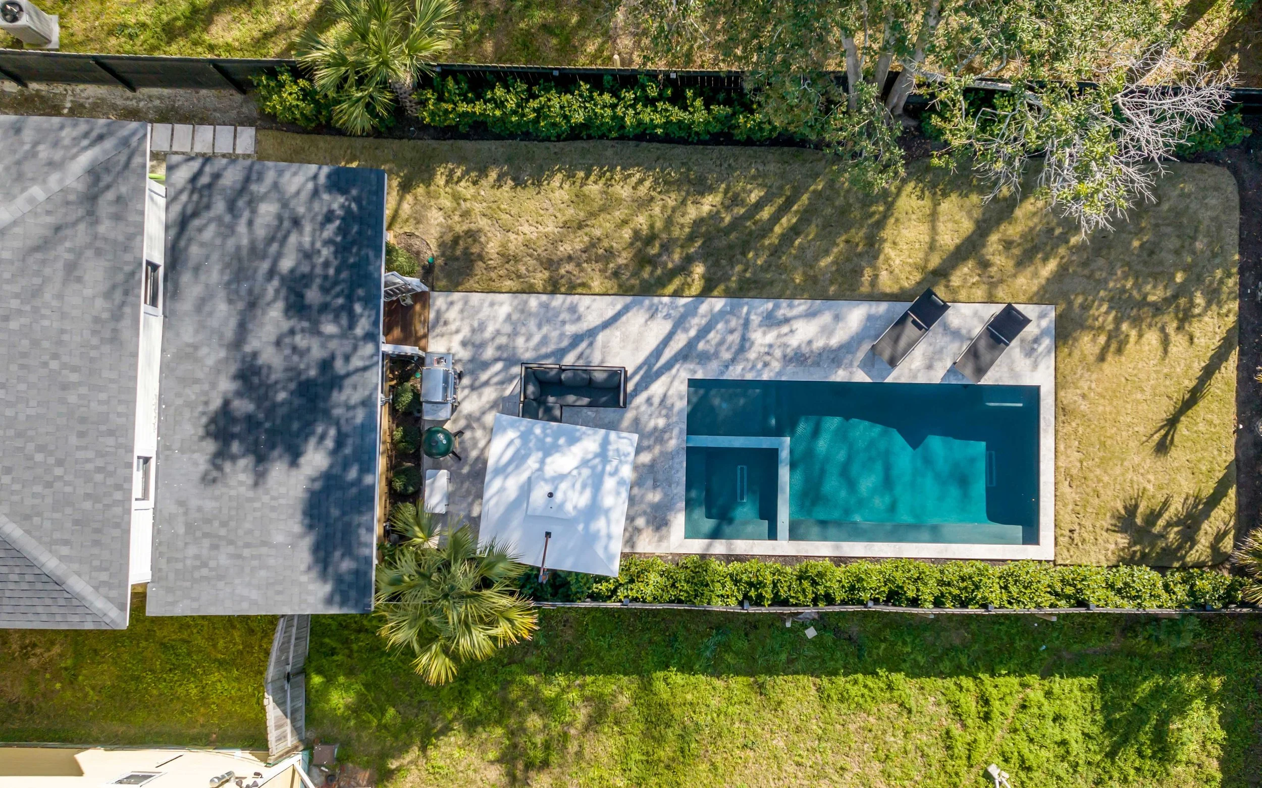 An aerial view of a backyard with a swimming pool, lounge chairs, a patio with outdoor furniture, a small garden area, and surrounding trees and grass.