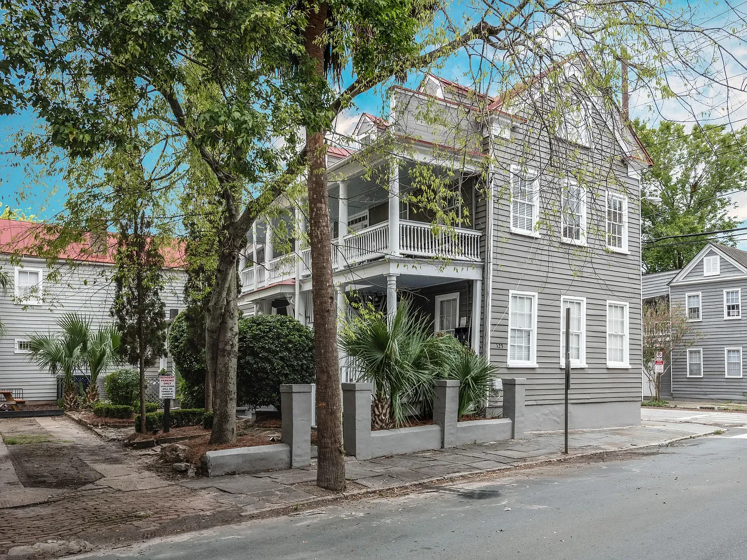 A gray, three-story residential building with white window frames and a front porch with white railings, surrounded by trees and greenery, located on a city street.