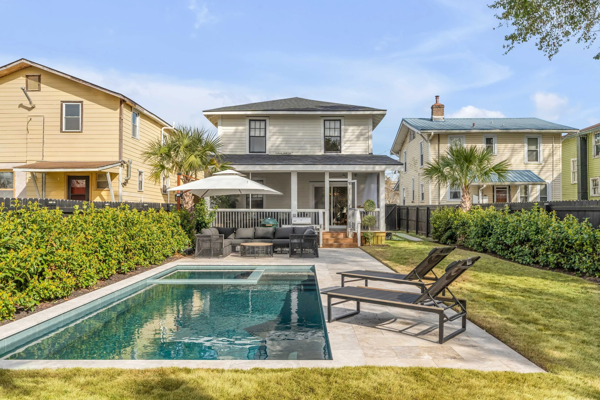 Backyard with swimming pool, two lounge chairs, patio furniture, umbrella, grass, bushes, and houses in the background.