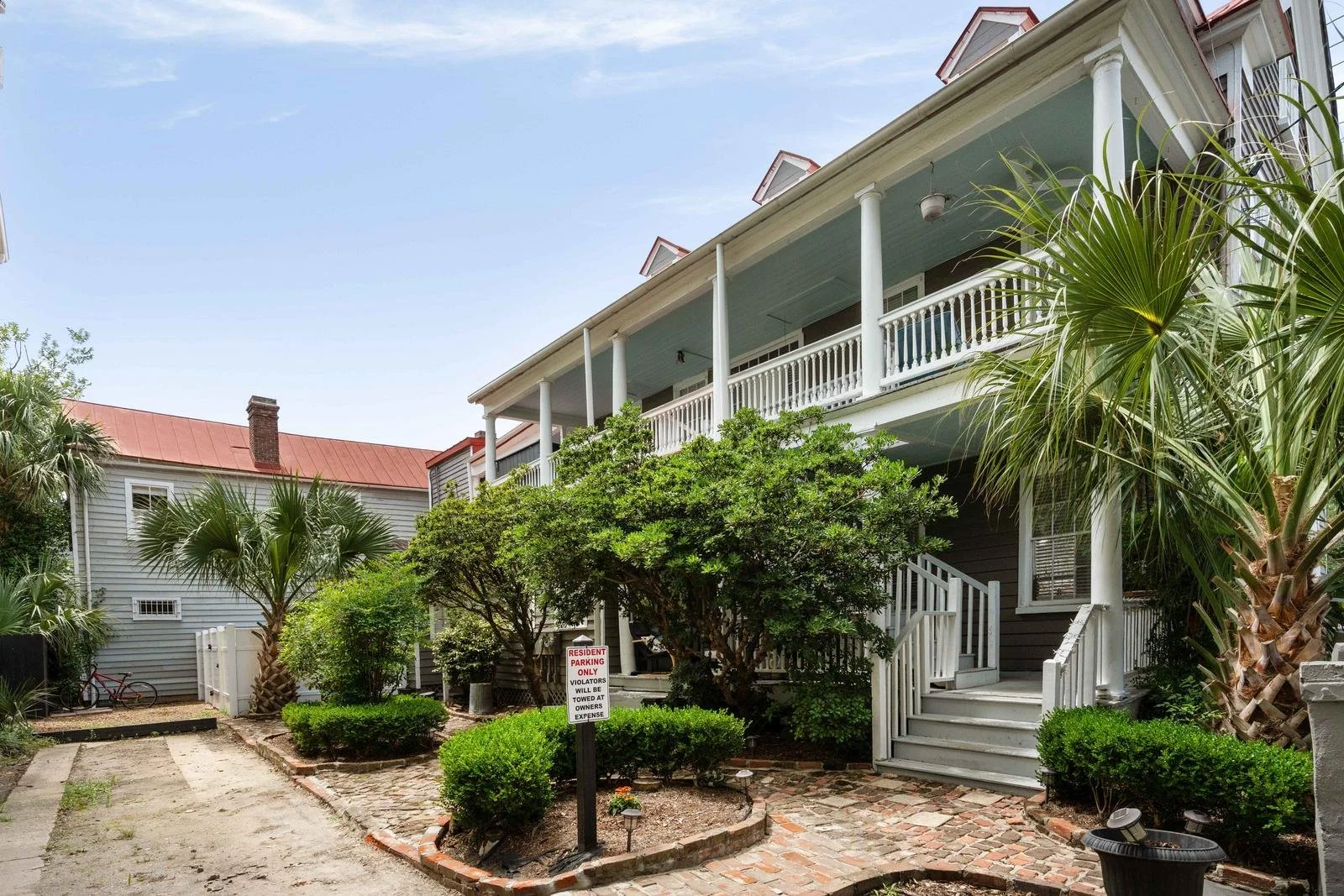 Two-story residential building with a large balcony, surrounded by lush green bushes and palm trees, with a brick-paved pathway and a parking area.