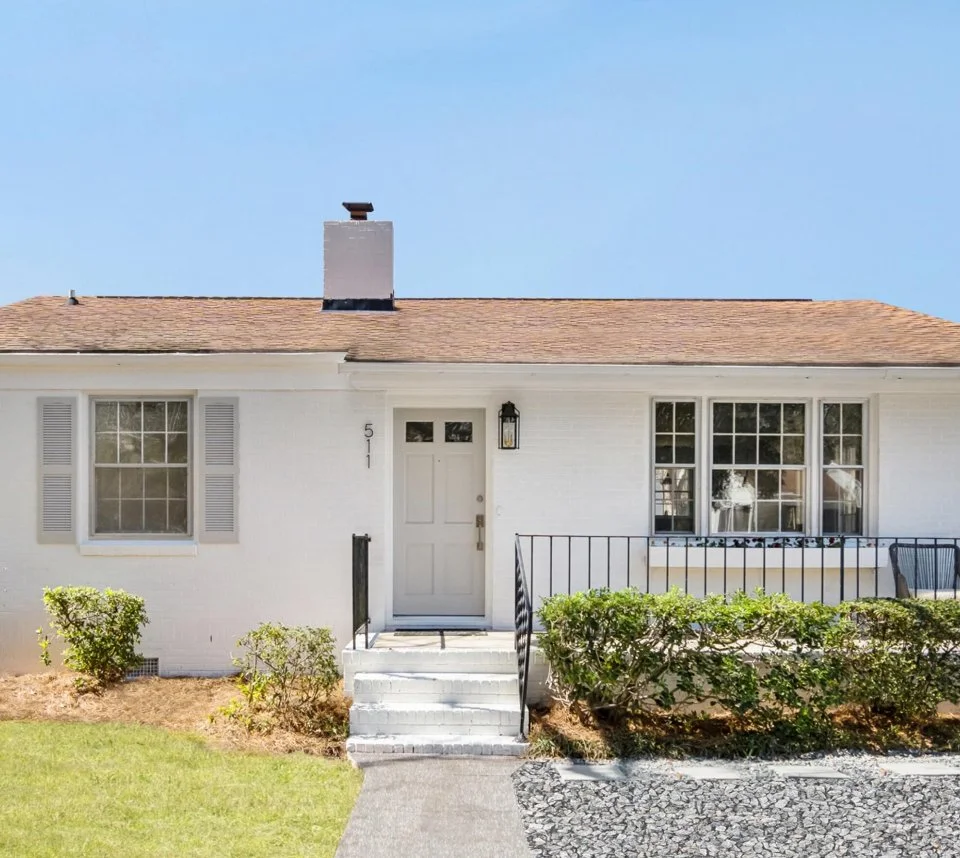Front view of a white single-family house with a brown shingle roof, a chimney, and large front windows. The house has a white door with a black lantern-style light fixture beside it, small steps leading up to the door, and a small front yard with bu