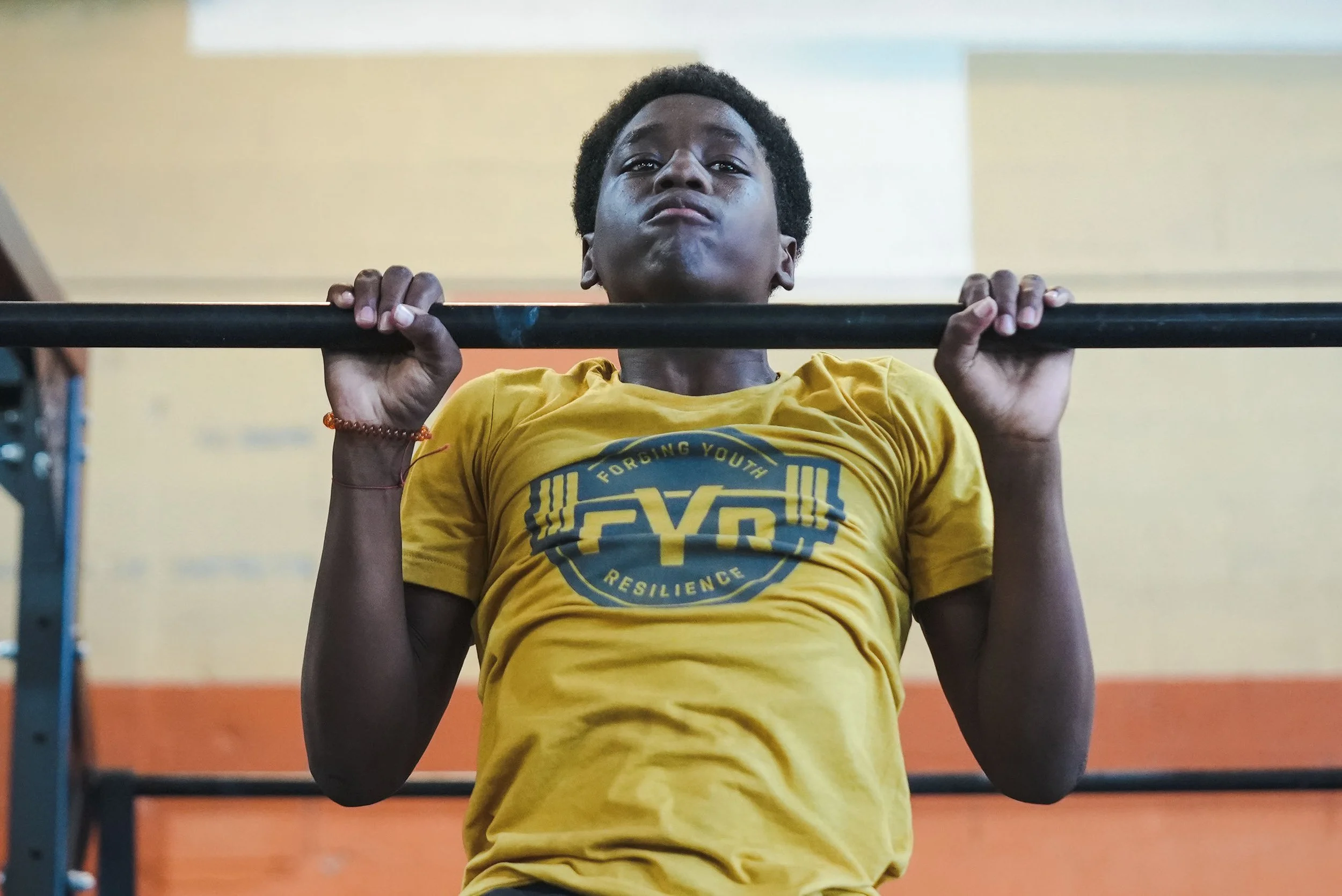 A young man with curly hair performing a pull-up exercise while holding a horizontal bar, wearing a yellow T-shirt with a logo that reads "Forcing Youth Resilience."