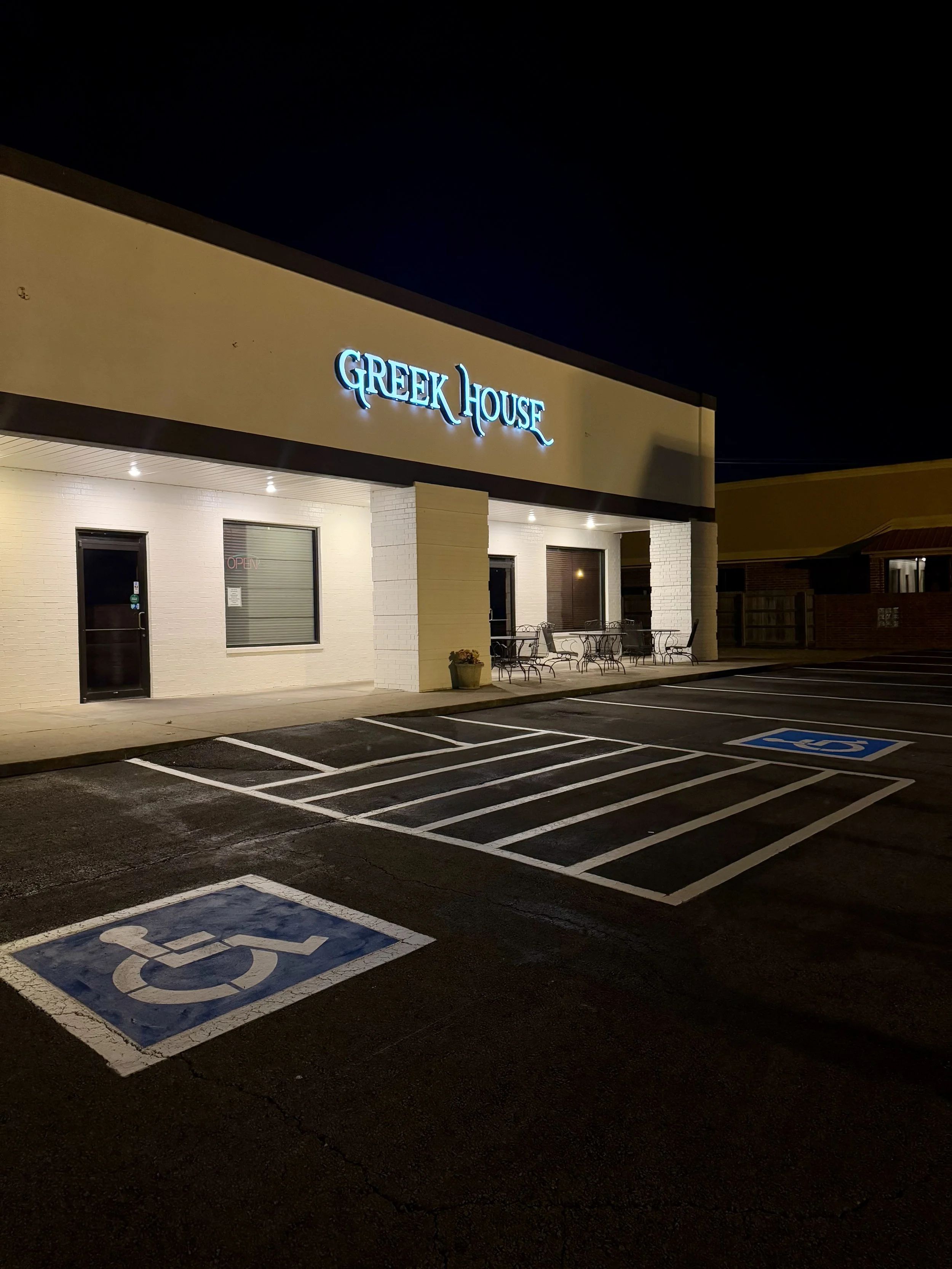 Night view of a Greek House restaurant with parking spaces, including handicapped spots, in the foreground. The building has a lit sign reading 'GREEK HOUSE' and outdoor seating with tables and chairs.