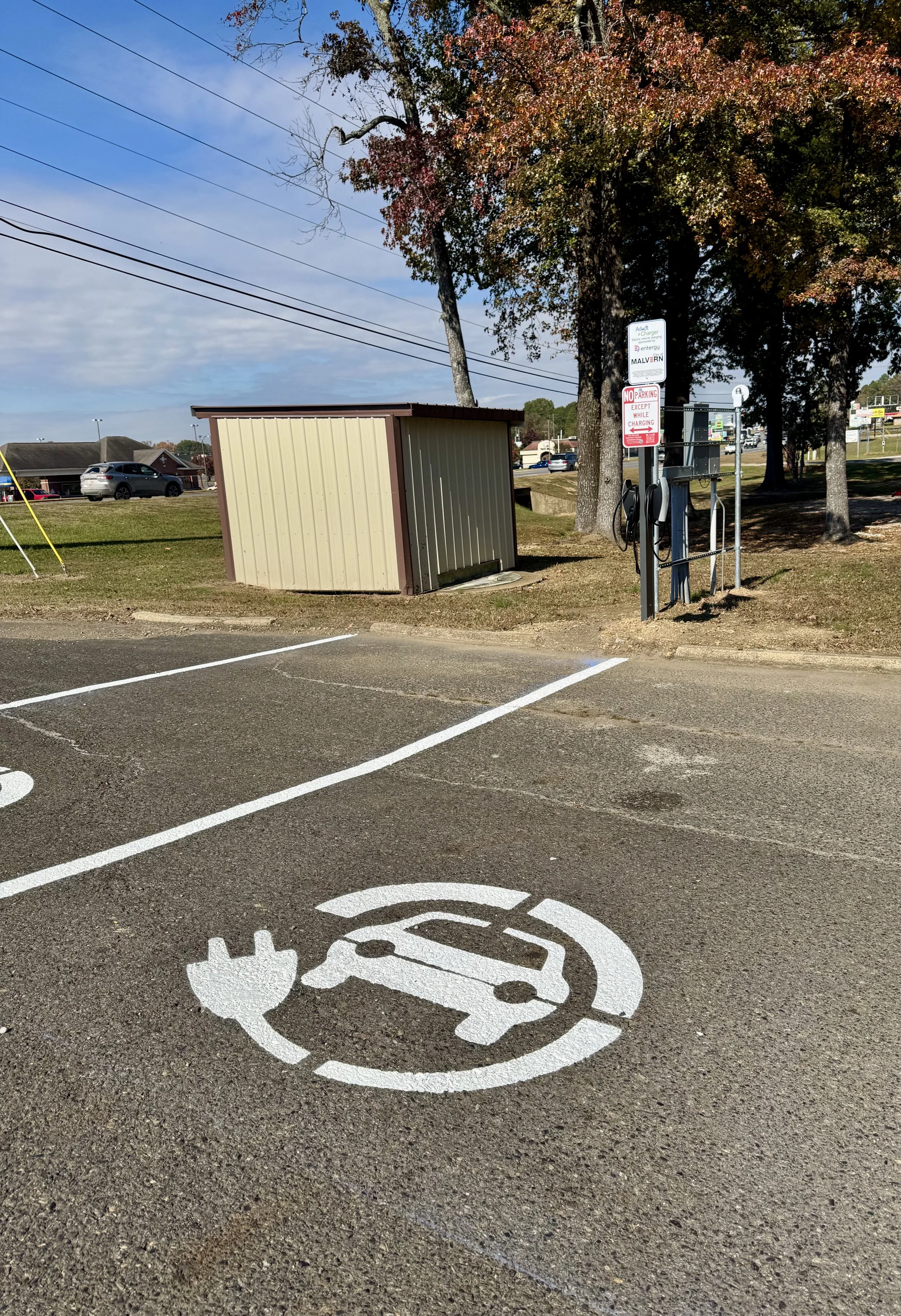 Electric vehicle charging station painted on parking lot asphalt with a symbol of a car and a plug. In the background, there is a small beige shed, trees with autumn foliage, and several signs including parking regulations.