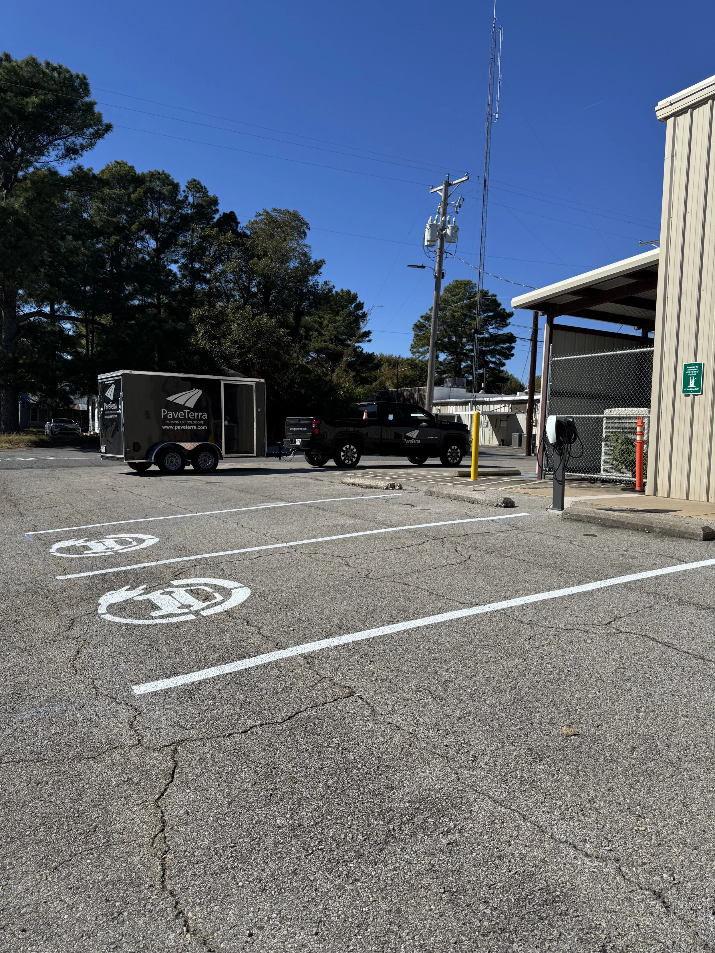 A parking lot with three designated parking spaces, two of which have white circle markings with broken lines, and the third one marked with a large white 'EV' for electric vehicle. There is a black pickup truck parked, a trailer hitched to it, and a