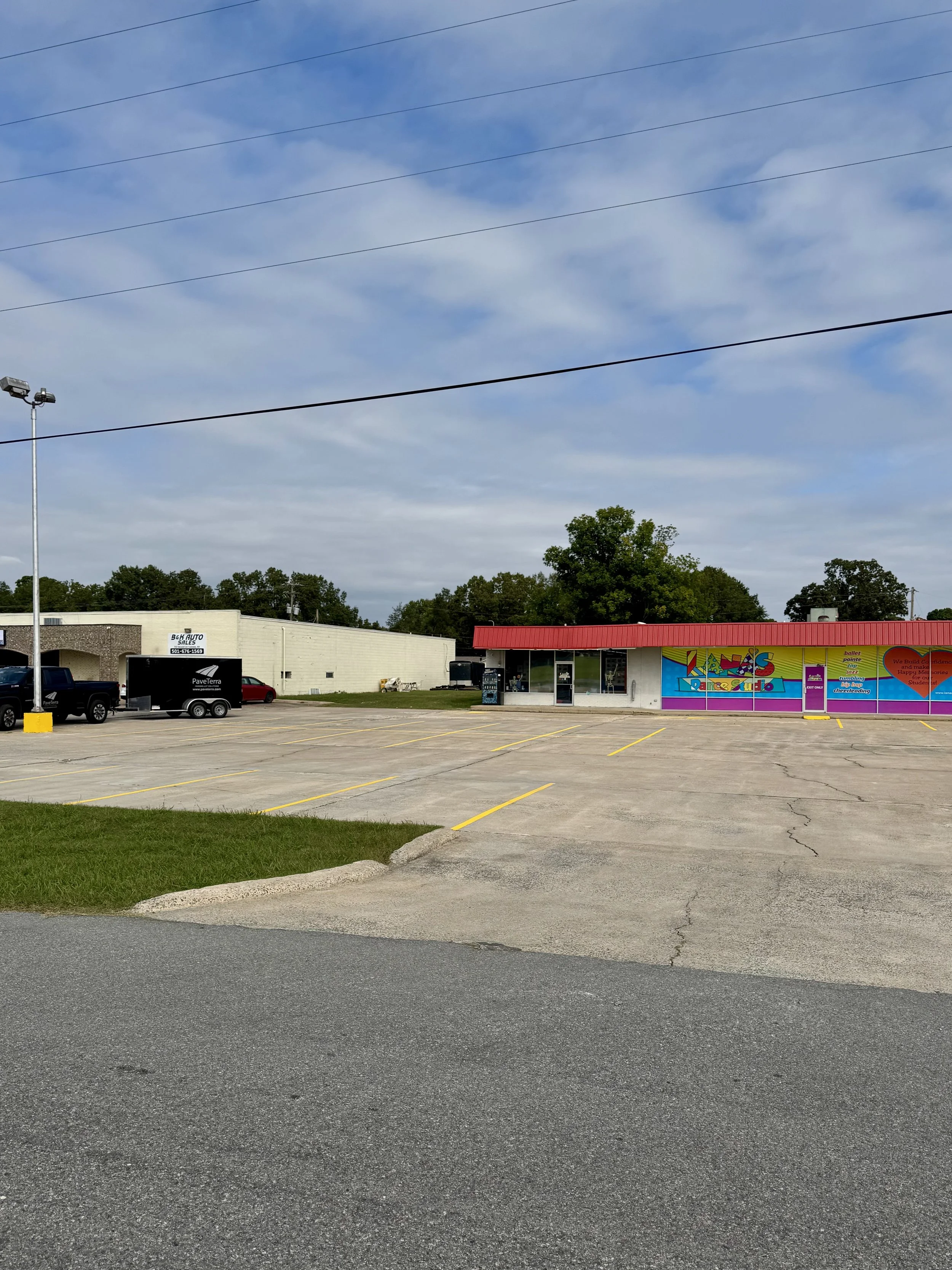 A small shopping plaza with a store called Kids R Kids daycare and learning center, a parking lot with a few cars, and a colorful mural on the building next to the daycare, under a partly cloudy sky.