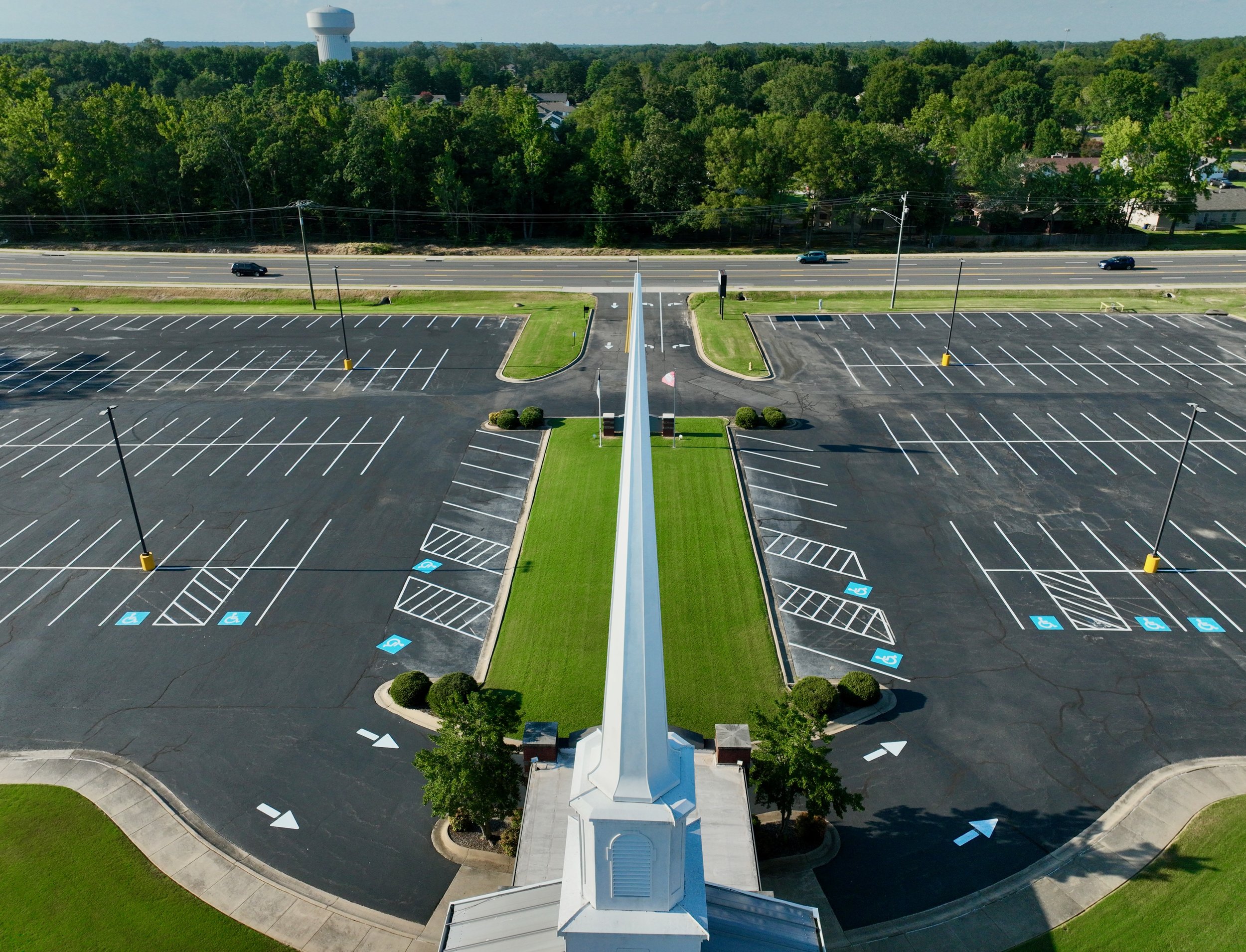 Empty parking lot with designated handicapped spaces, marked with blue and white signs. No cars parked, view from above with church steeple in the foreground and a forested area and water tower in the background.